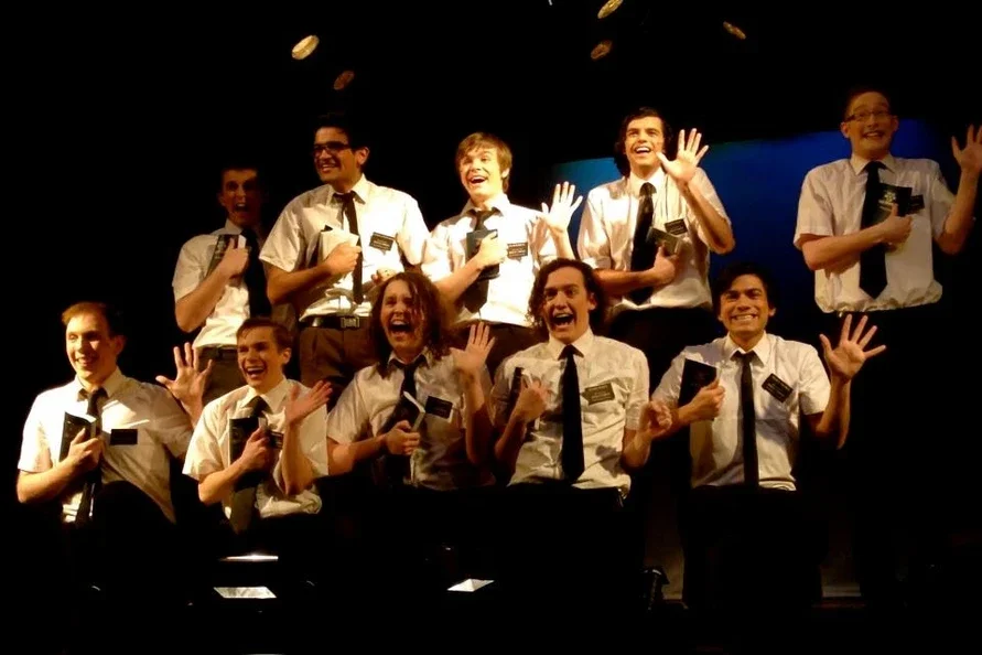 Group of young men wearing white shirts, black ties, and glasses on stage, smiling and waving holding Books of Mormon