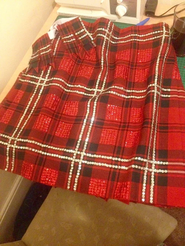 A red tartan kilt with a pattern of silver sequins arranged in lines, laid out on a work table with a green cutting mat, sewing tools, and a machine in the background.