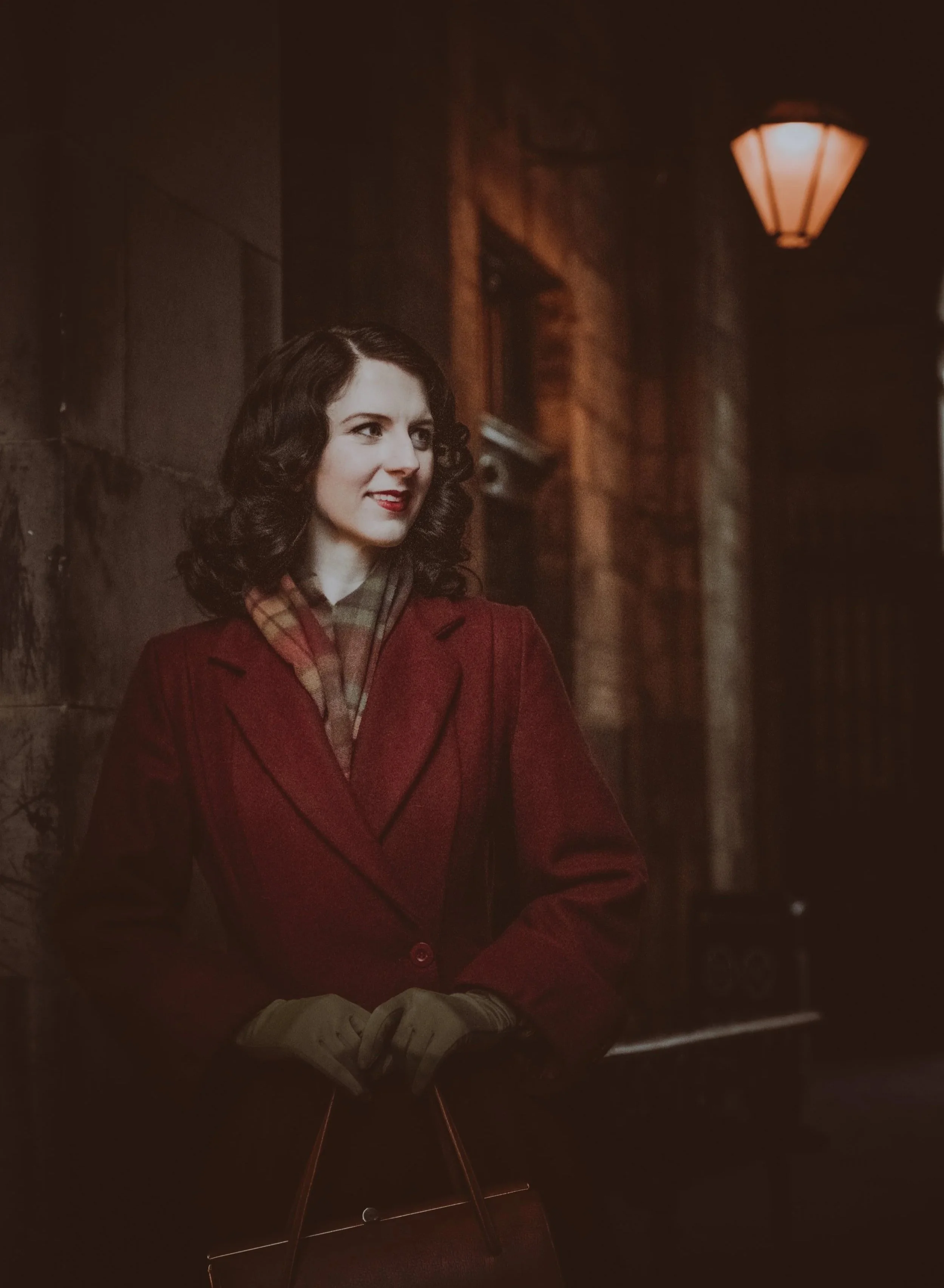 A woman with dark wavy hair, wearing a red wool coat and an autumnal tartan scarf, standing outside at night near a stone wall and an old fashioned lamp. She is smiling and looking to her right.