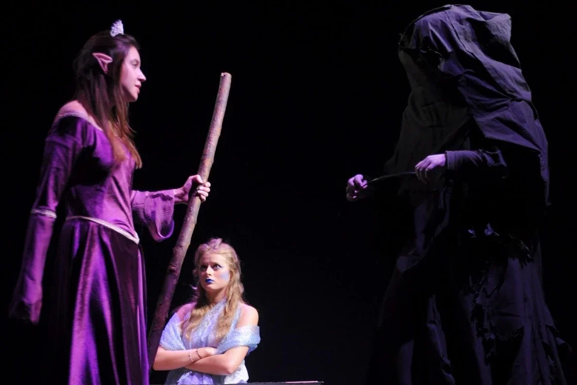 Three actors on stage during a theatrical performance. A woman dressed as a witch in a purple costume holding a staff, a young girl with blonde hair and blue makeup looking up with crossed arms, and a figure dressed in black with a hooded cloak conce