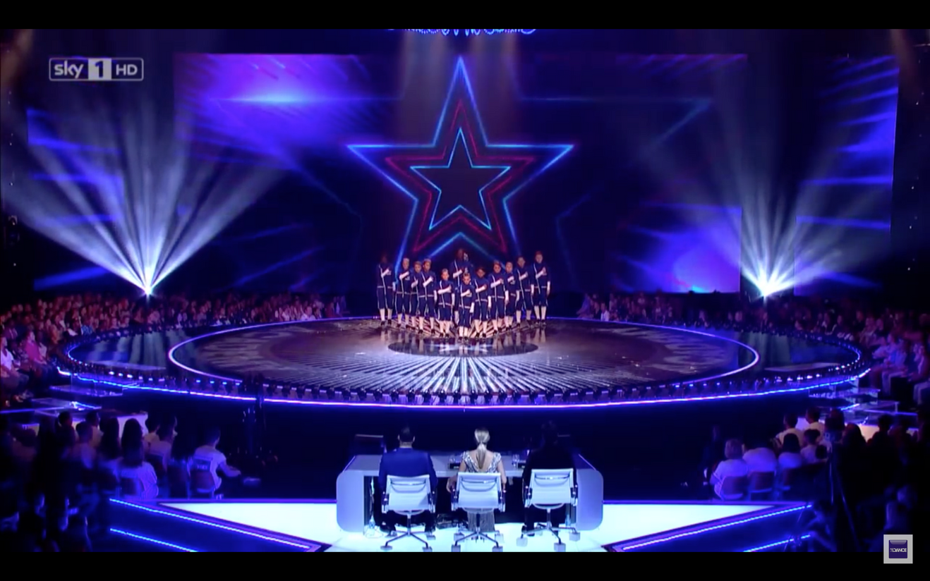 A group of dancers standing in a large circular stage with blue and purple lighting. A giant star-shaped neon light is projected on a screen behind them, and there is an audience seated around the stage.