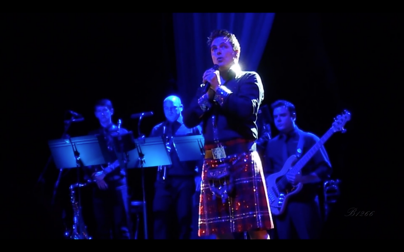 John Barrowman performing on stage in a sequinned kilt with bandmates in the background, illuminated by stage lights.