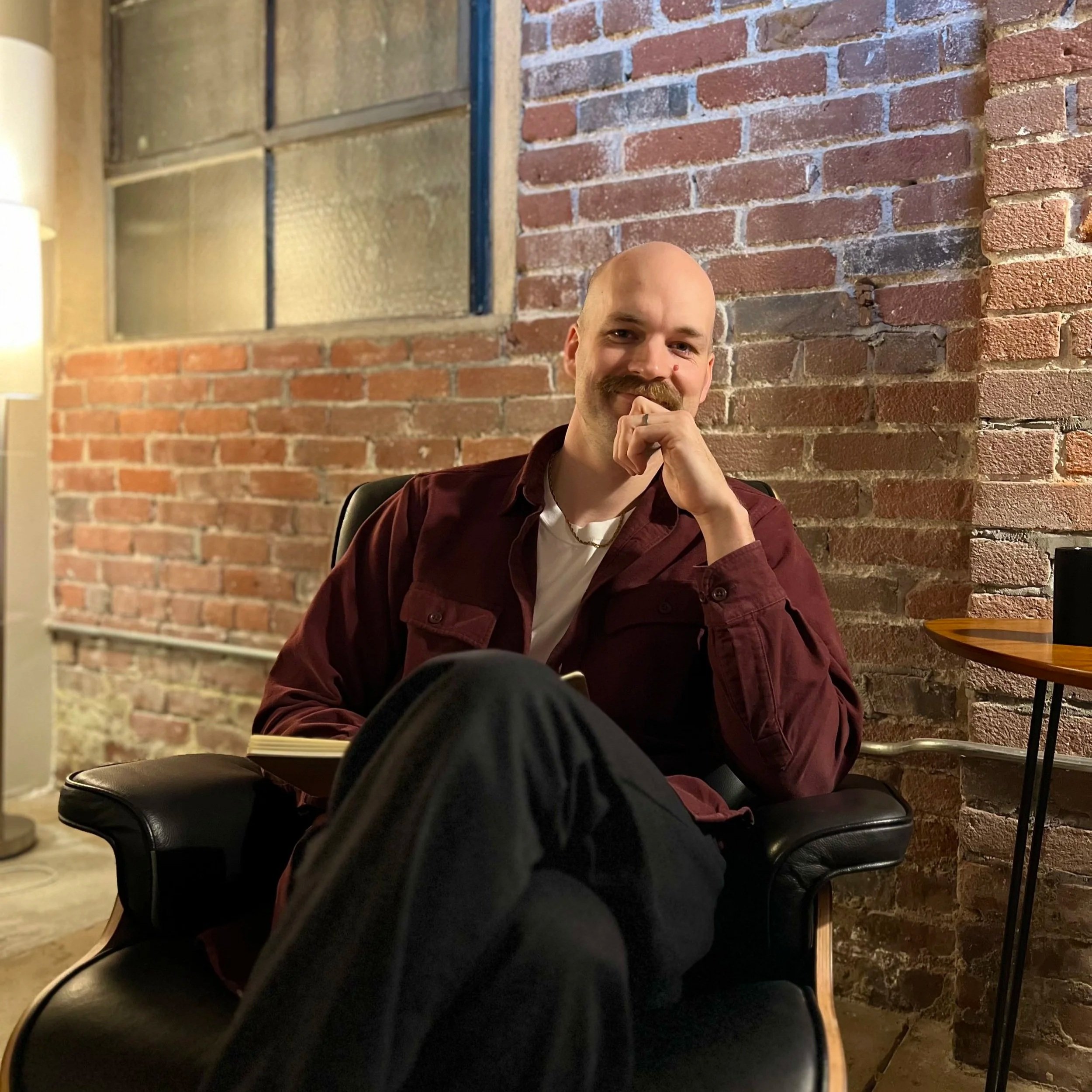 A man with a mustache and bald head sitting indoors in front of an exposed brick wall, wearing a maroon jacket and white t-shirt, resting his chin on his hand.