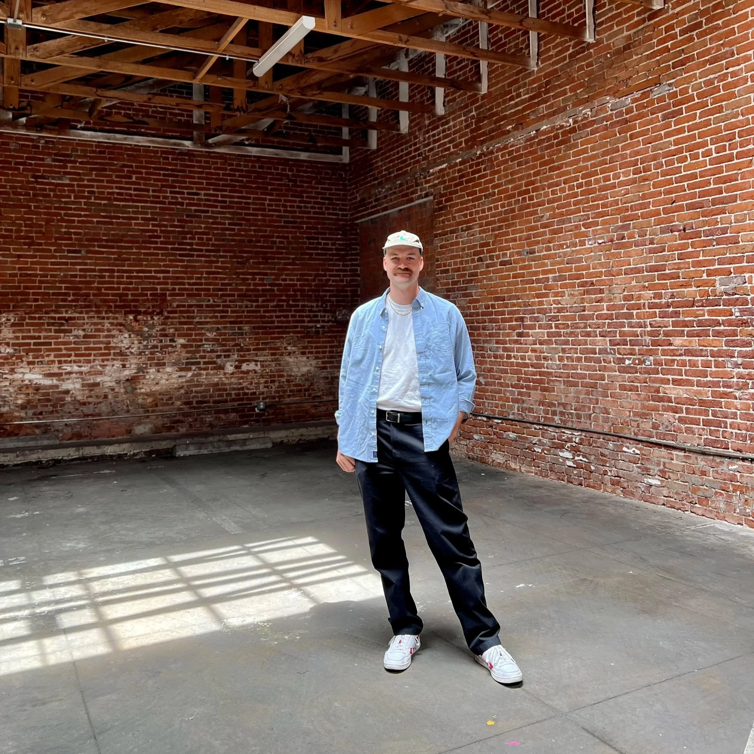 A man wearing a light blue shirt, white t-shirt, dark pants, white sneakers, and a white cap standing against a brick wall with shadows cast by window blinds.
