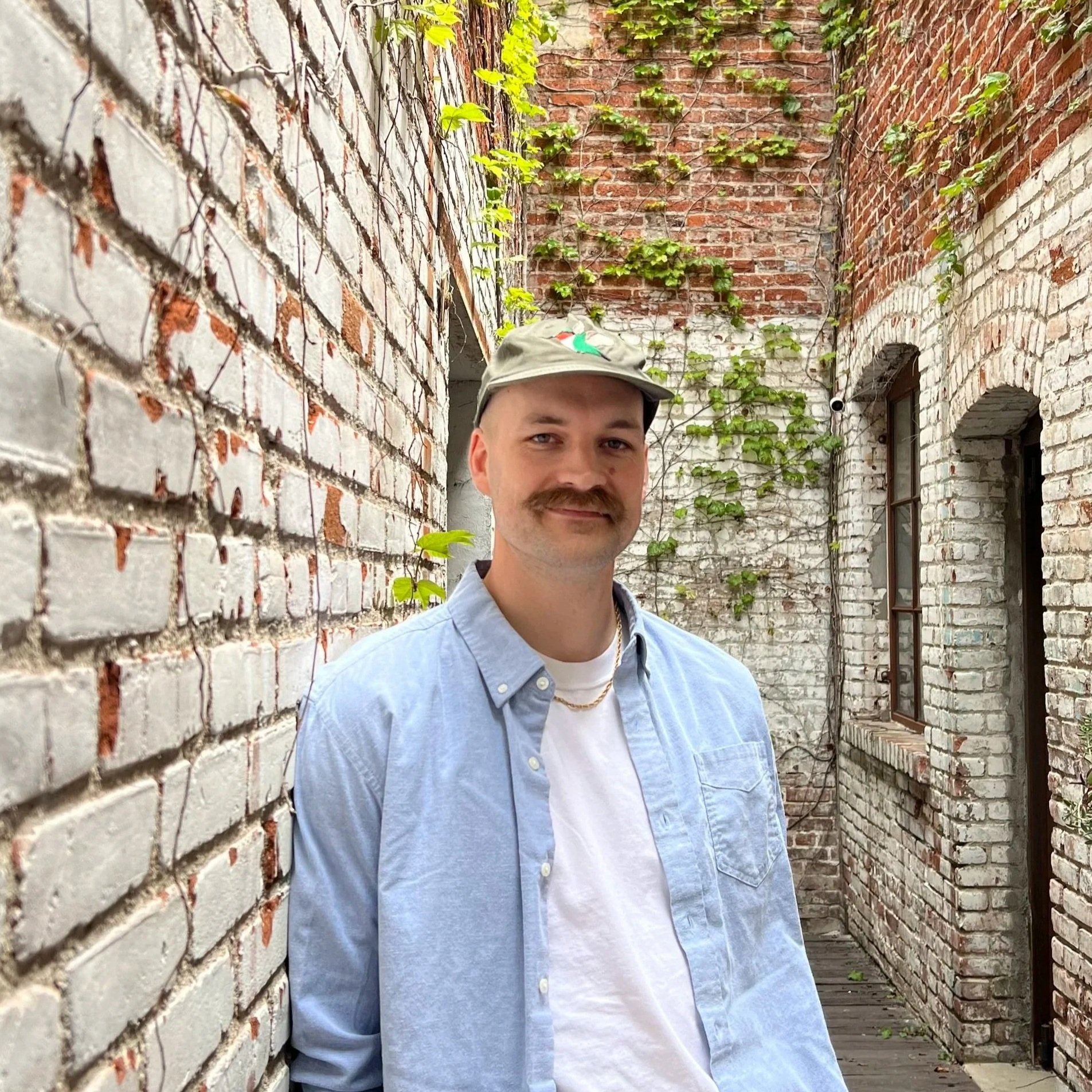 A young man with a light blue button-up shirt, a gold chain necklace, and a khaki baseball cap with a colorful bird embroidered on it, standing in an alley between brick walls covered with vines and greenery.