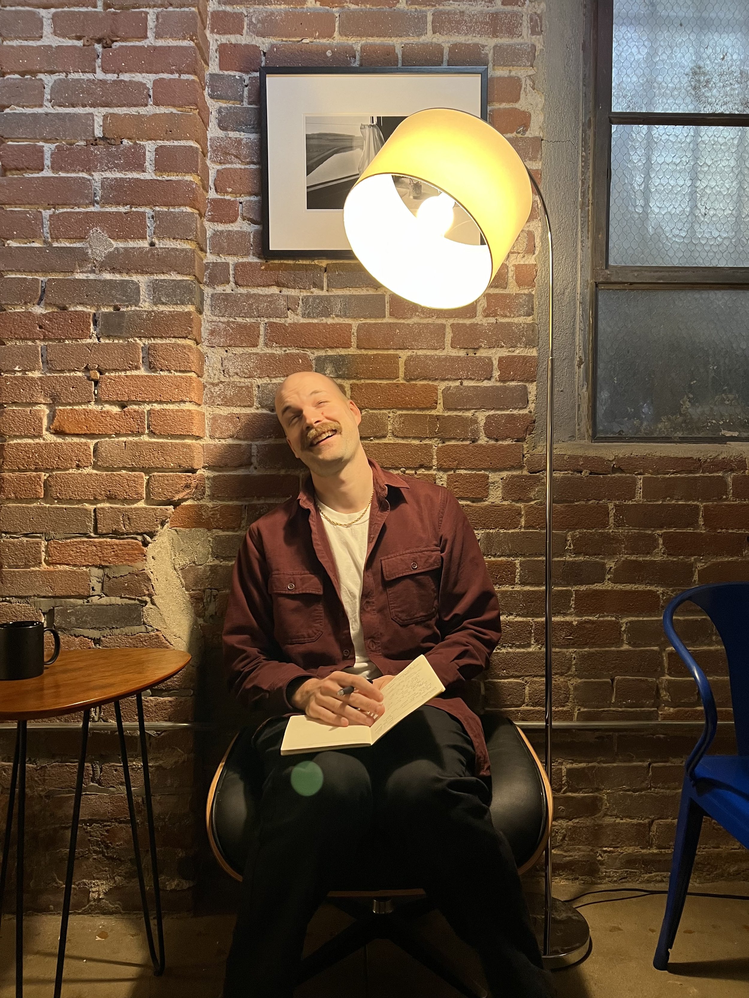 A man with a mustache and a goatee, wearing a maroon jacket over a white shirt, sitting on a black stool in front of a brick wall with a framed black and white photo, holding an open notebook and pen, smiling.