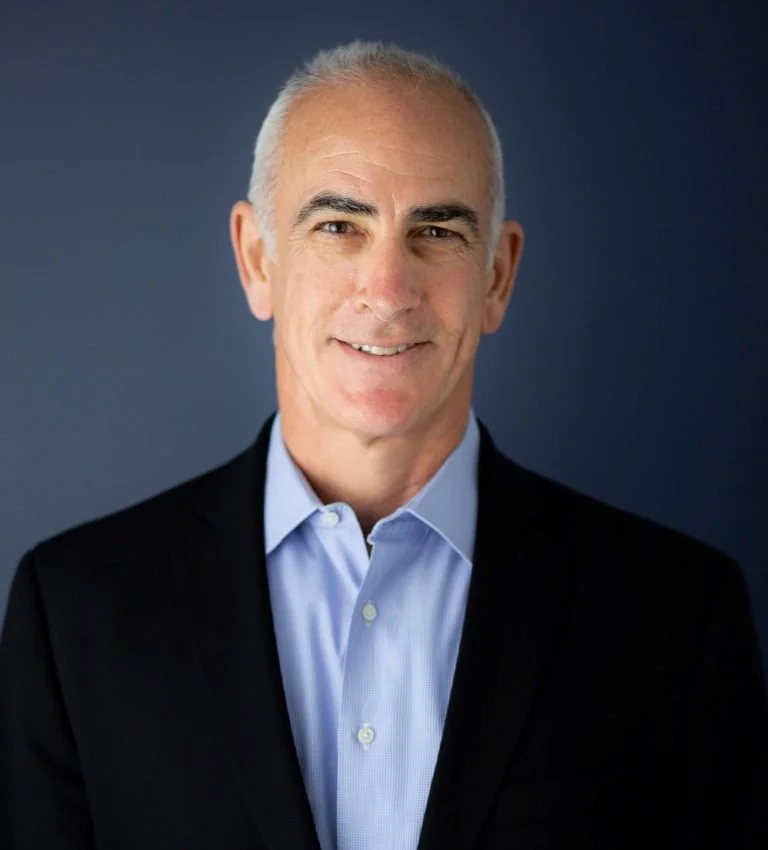 A middle-aged man with short silver hair, wearing a dark suit jacket and a light blue dress shirt, standing against a dark background and smiling at the camera.