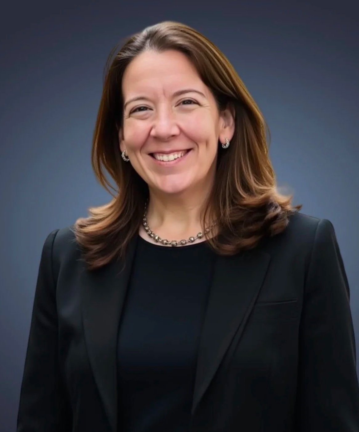 A smiling woman with brown hair in loose waves, wearing a black blazer, a pearl necklace, and pearl earrings, standing against a plain dark background.