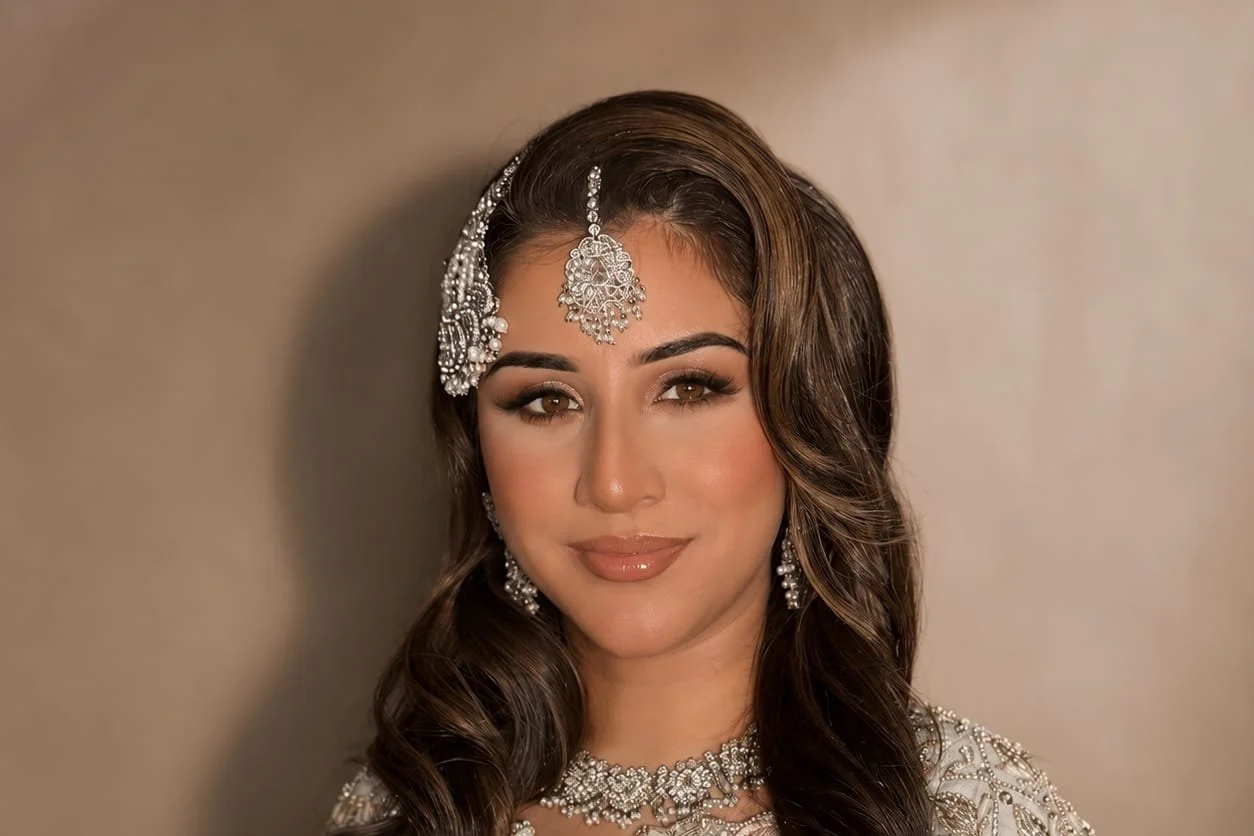 A woman with long, wavy brown hair wearing traditional Indian jewelry and attire, including a headpiece, earrings, necklace, and matching dress, with makeup and a neutral background.
