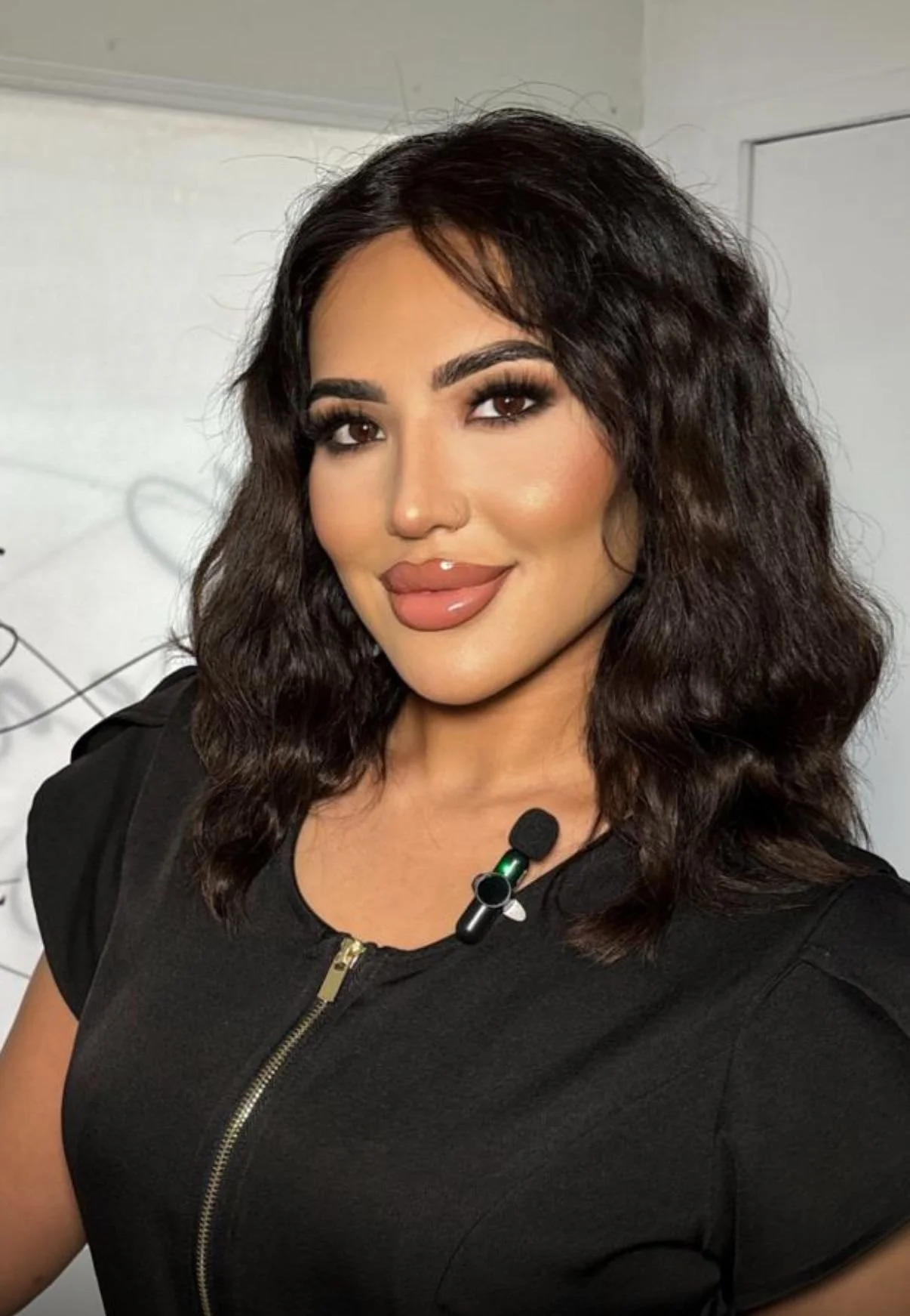 A woman with dark wavy hair and makeup, wearing a black top with a zipper, and a microphone clipped to her shirt, standing in front of a light-colored wall.