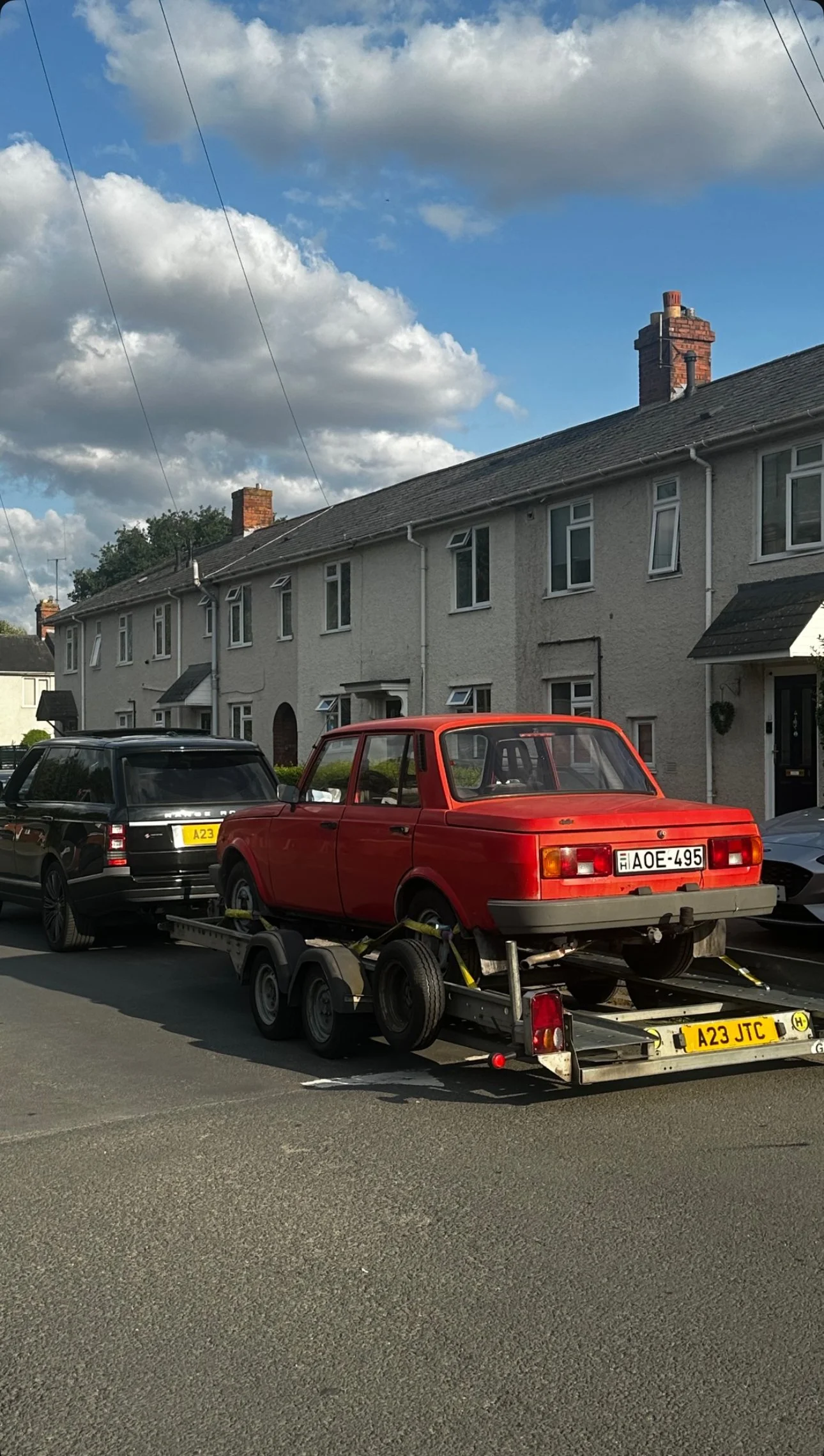 A red vintage car is on a trailer being towed on the street, with a black modern car parked behind it. The background features residential houses with white walls and chimneys, and a partly cloudy sky.