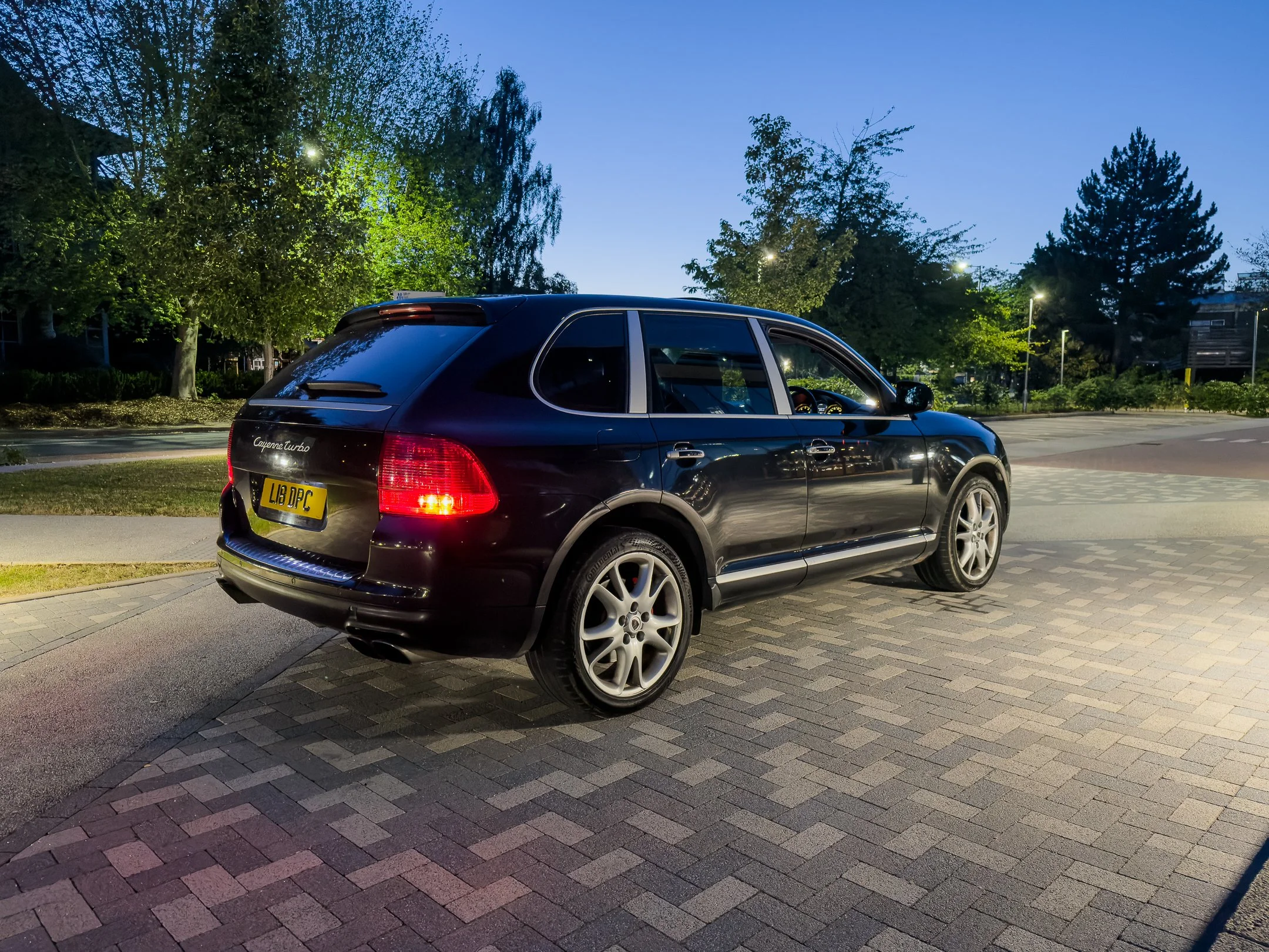 A black Porsche Cayenne Turbo parked on a paved area under a canopied street lights with trees and dark blue sky in the background.