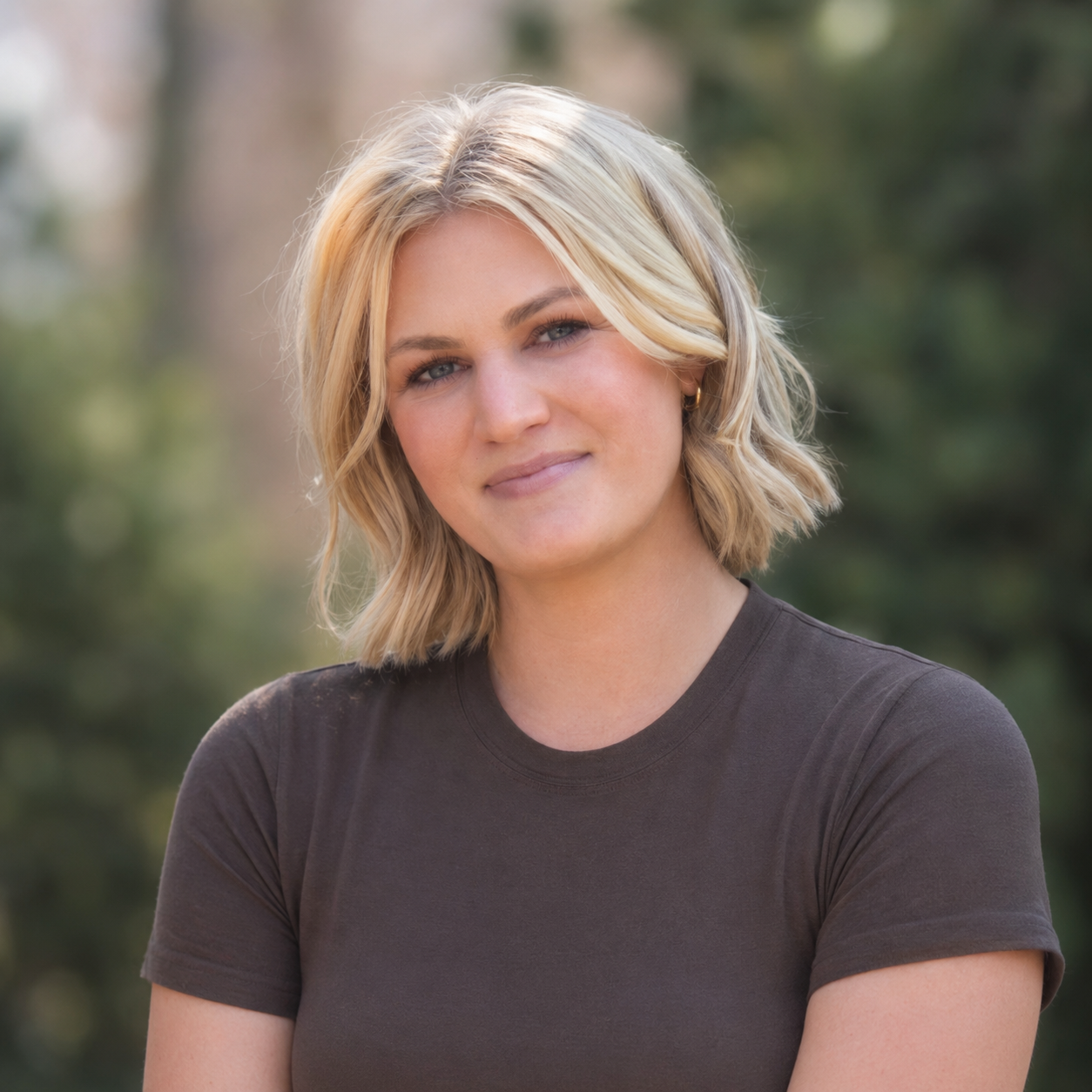 A woman with shoulder-length blonde hair and a slight smile, wearing a black t-shirt, outdoors with blurred green trees in the background.