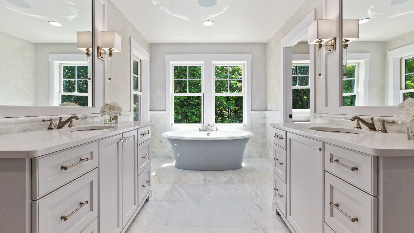 A bright, spacious bathroom with a freestanding bathtub in front of a large window, double vanity sinks on either side, and mirrors with wall-mounted lights, all in a clean, white decor style.