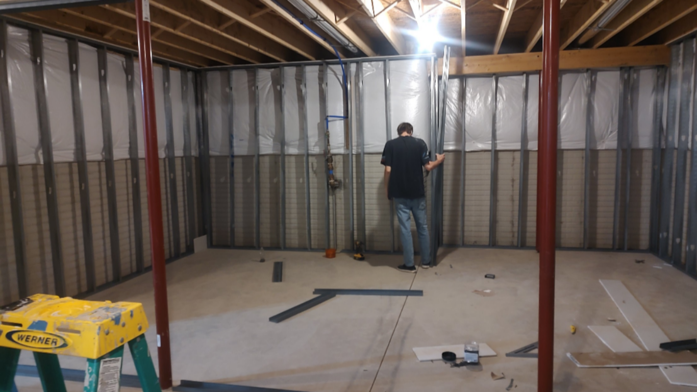 Basement remodeling and wall framing process with steel studs, insulation vapor barrier, step ladder, and a contractor building out a basement room.