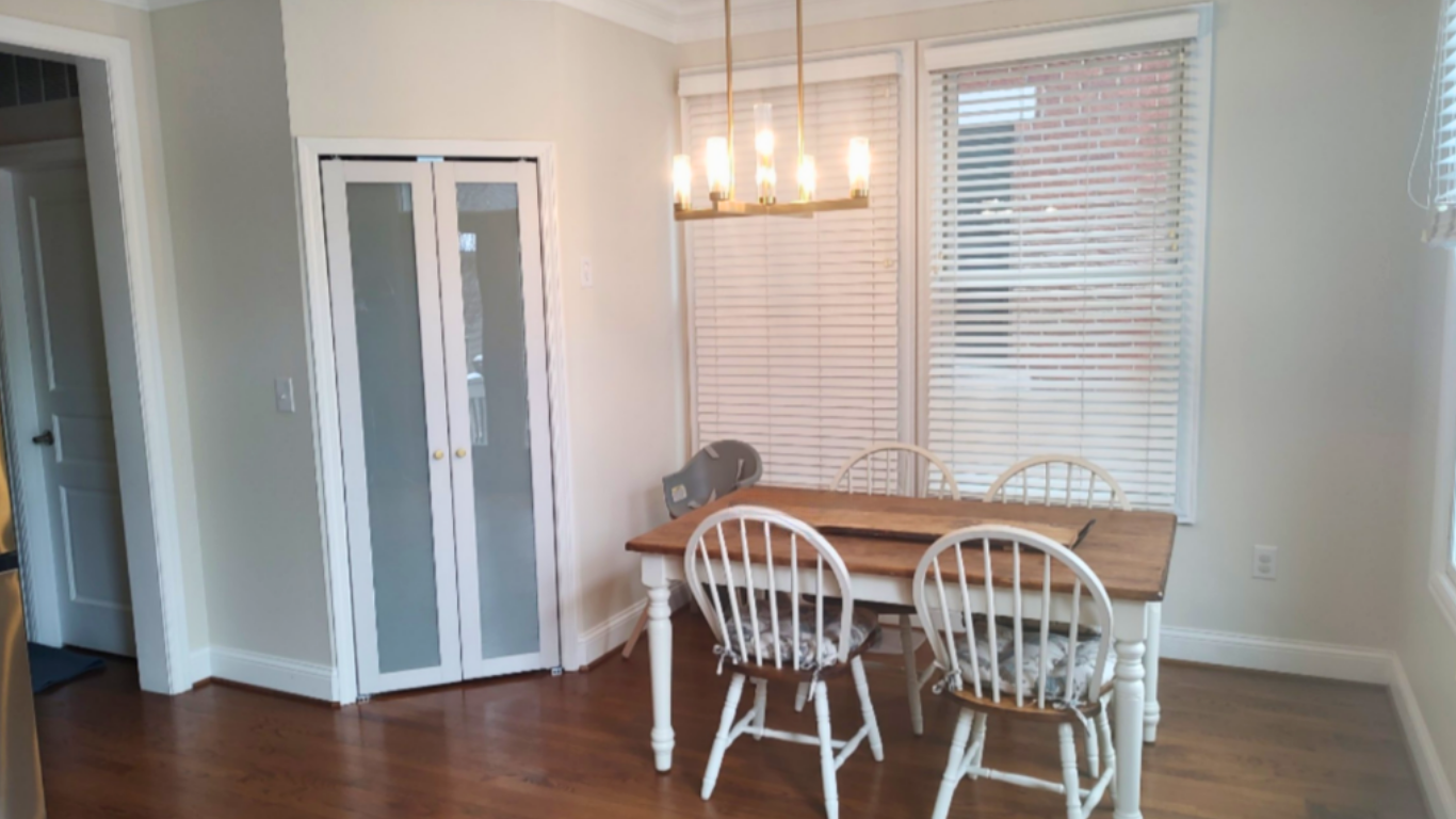 A dining area with a wooden table, four white chairs with patterned cushions, large windows with white blinds, a modern chandelier with multiple exposed bulbs, and a doorway with frosted glass double doors.
