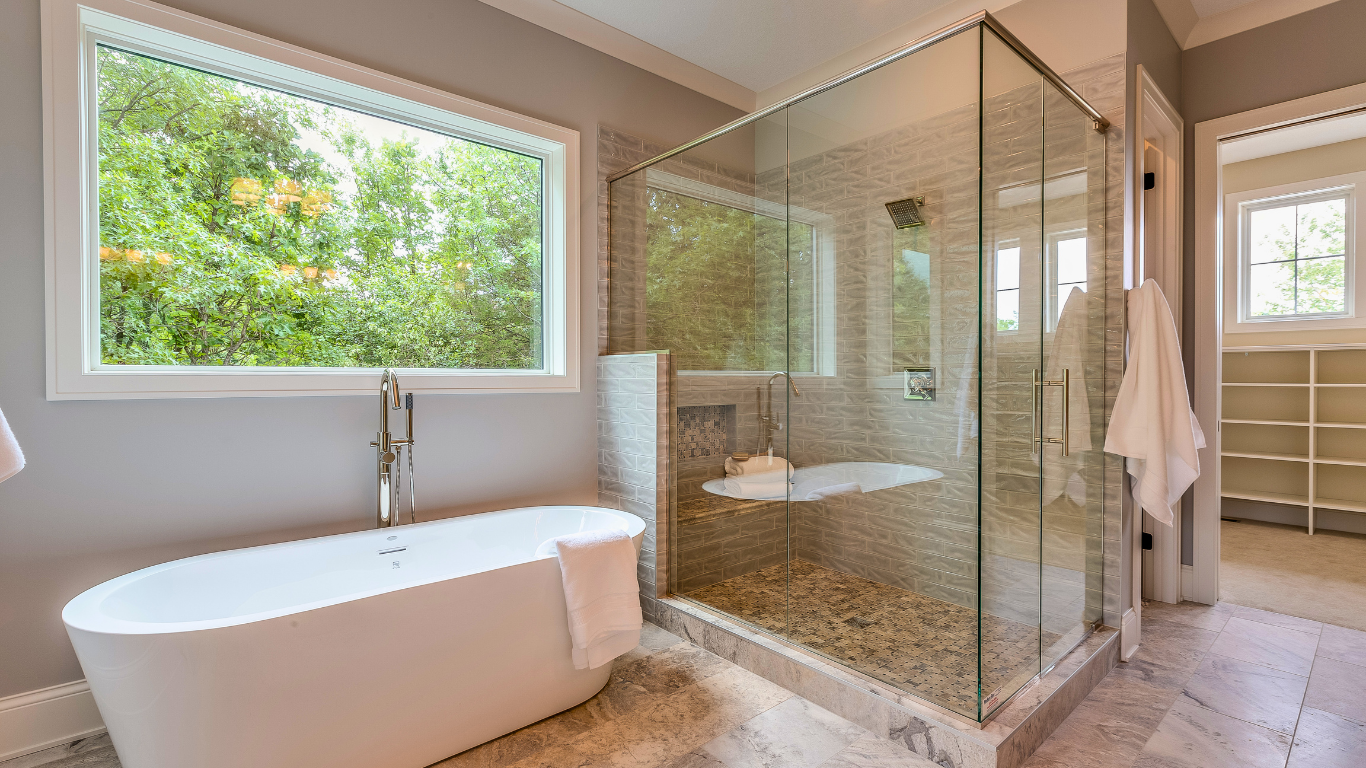 Bright bathroom with a large window overlooking trees, featuring a white freestanding soaking tub and a corner glass-enclosed walk-in shower with stone tile flooring.
