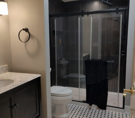 A bathroom featuring a walk-in shower with vertical dark charcoal tiles and a black-framed sliding glass door, paired with a white toilet and a black and white basketweave mosaic tile floor.