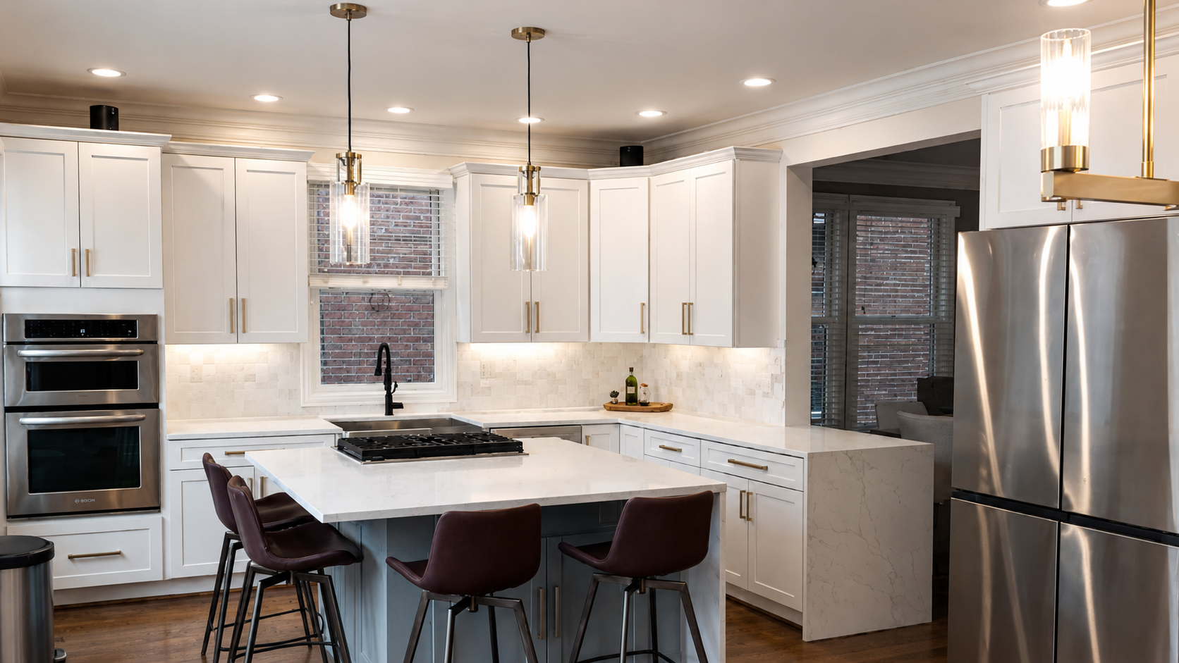 Modern white kitchen with stainless steel appliances, including a double oven and refrigerator, a kitchen island with four brown chairs, pendant lights, and a window showing brick wall outside.