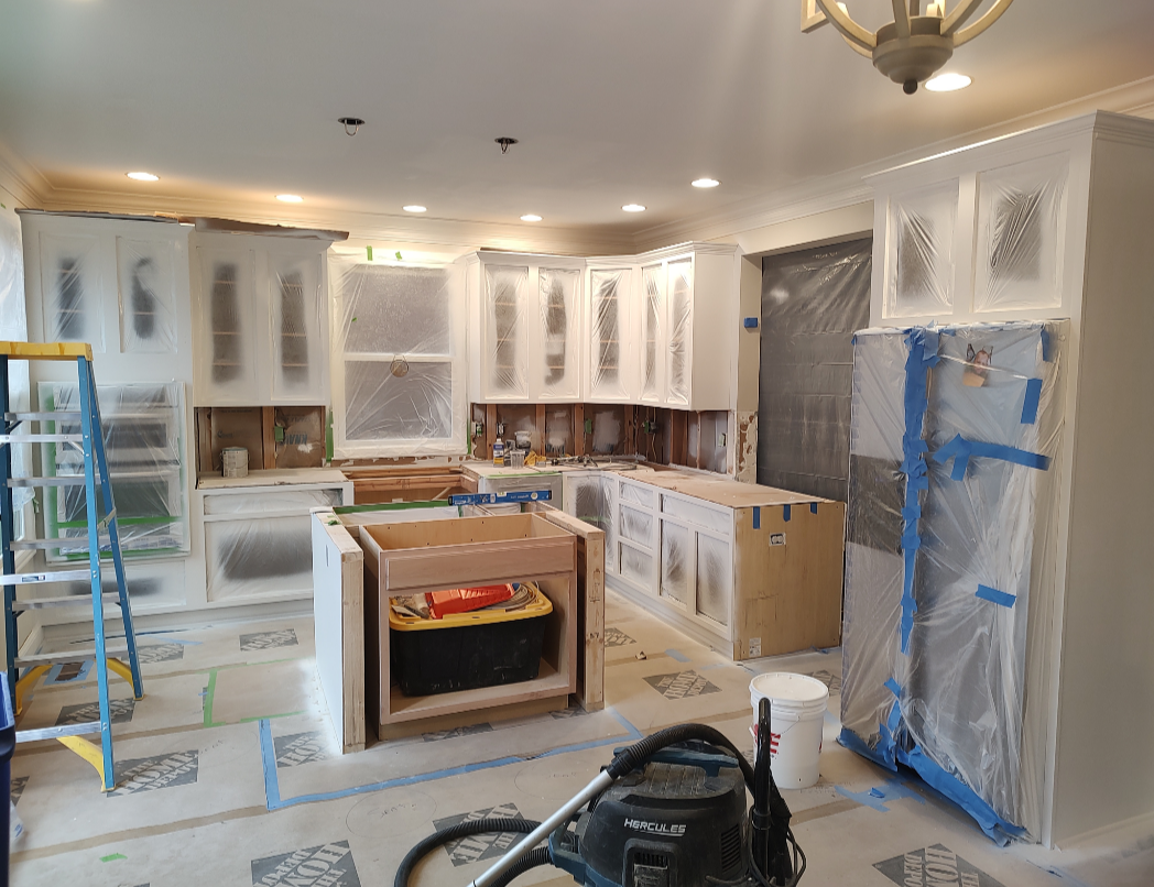 Kitchen cabinet refacing and painting process with masked doors, protective plastic sheets, shop vac, and step ladder during a home remodel.