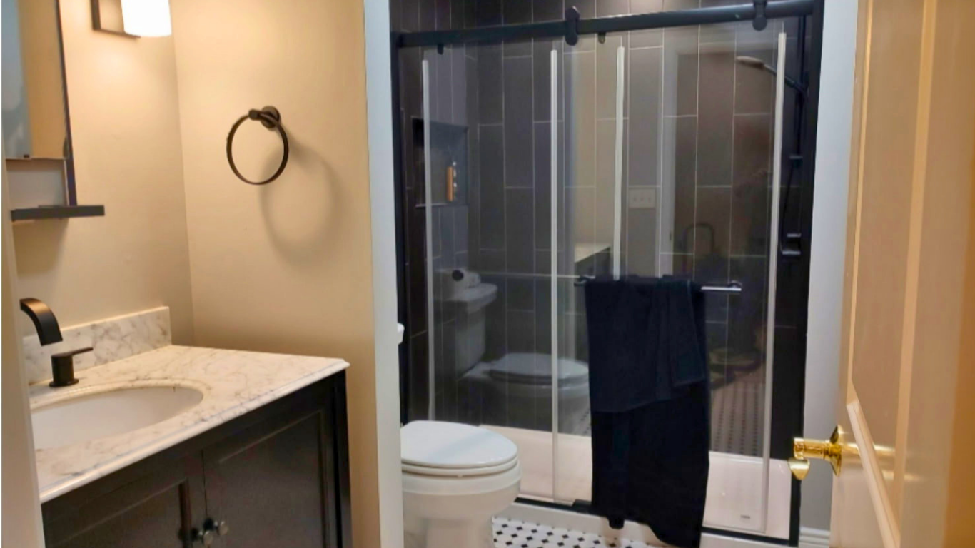 Bathroom with a vanity sink, marble countertop, black faucet, wall-mounted black towel ring, toilet, and a shower enclosed by glass sliding doors with black trim. The shower has gray tiles and a towel hanging on the door handle.