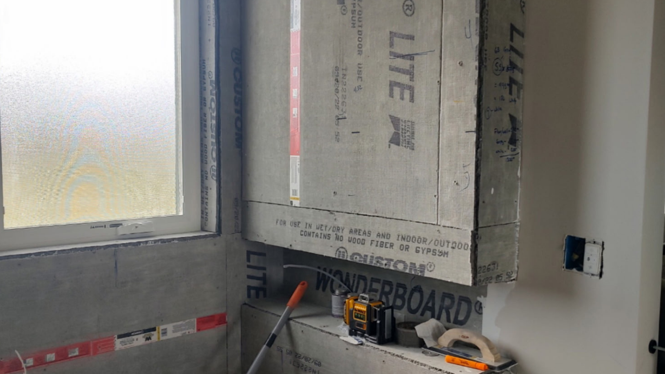 Bathtub tile preparation phase with cement backer board secured to the wall framing. Tile installation tools and a laser level sit on the unfinished bathtub ledge next to a frosted window during a bathroom remodel.