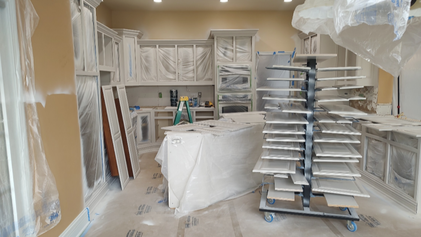 Kitchen cabinet refacing and painting process featuring taped-off cabinet boxes, freshly painted doors on a vertical drying rack, and plastic floor protection during a home remodel.