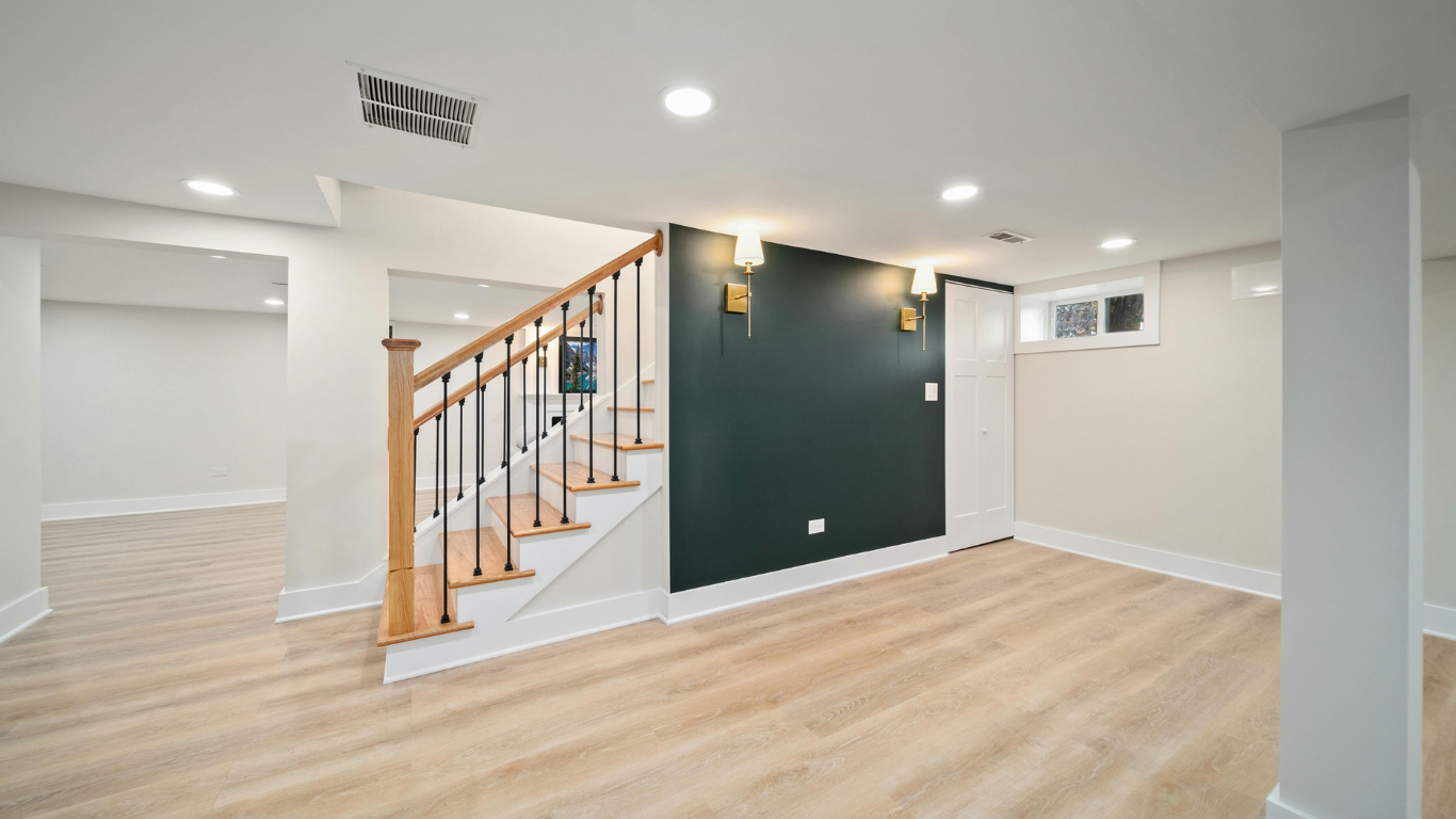 Brightly lit finished basement with light wood flooring, an open staircase with wooden treads, and a dark green accent wall with two gold sconces.