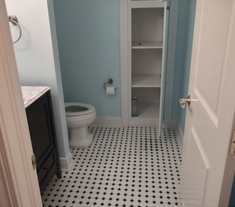 A small bathroom with light blue walls, a white built-in linen storage tower with open shelving, a white toilet, and a classic black and white basketweave mosaic tile floor.
