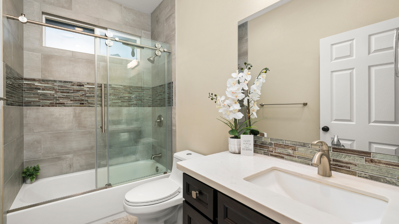 A modern, remodeled bathroom featuring a white bathtub with a sliding glass door enclosure, grey tiled walls with a mosaic stone accent strip, a white toilet, and a dark wood vanity with a white countertop and brushed nickel faucet.