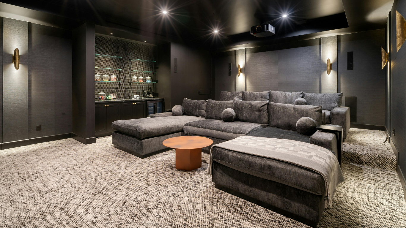 Dark home theater basement featuring a large charcoal grey sectional couch, patterned carpet, wall sconces, and a built-in snack bar in the background.