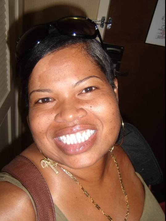 A woman smiling, wearing sunglasses on her head, gold jewelry, and a tank top.