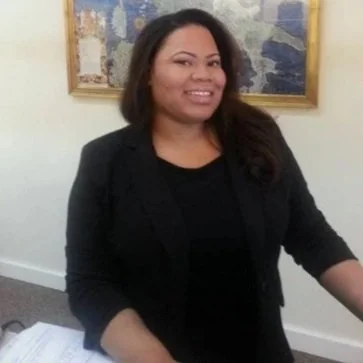 A woman with shoulder-length dark hair wearing a black blazer and black top, smiling, standing in front of an abstract painting.