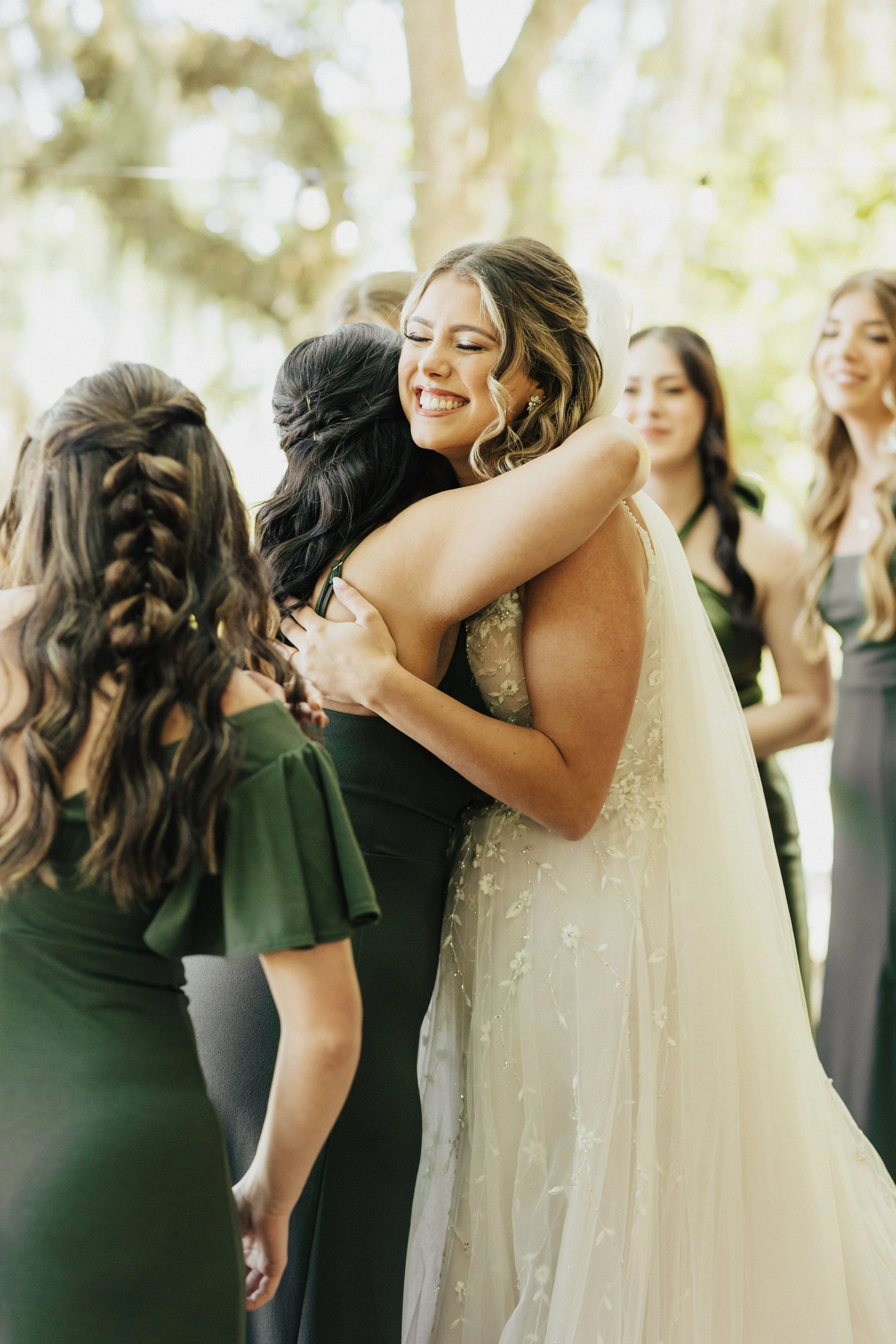A woman in a white wedding dress being hugged by another woman in a dark dress at an outdoor wedding ceremony, with other women in dresses in the background.