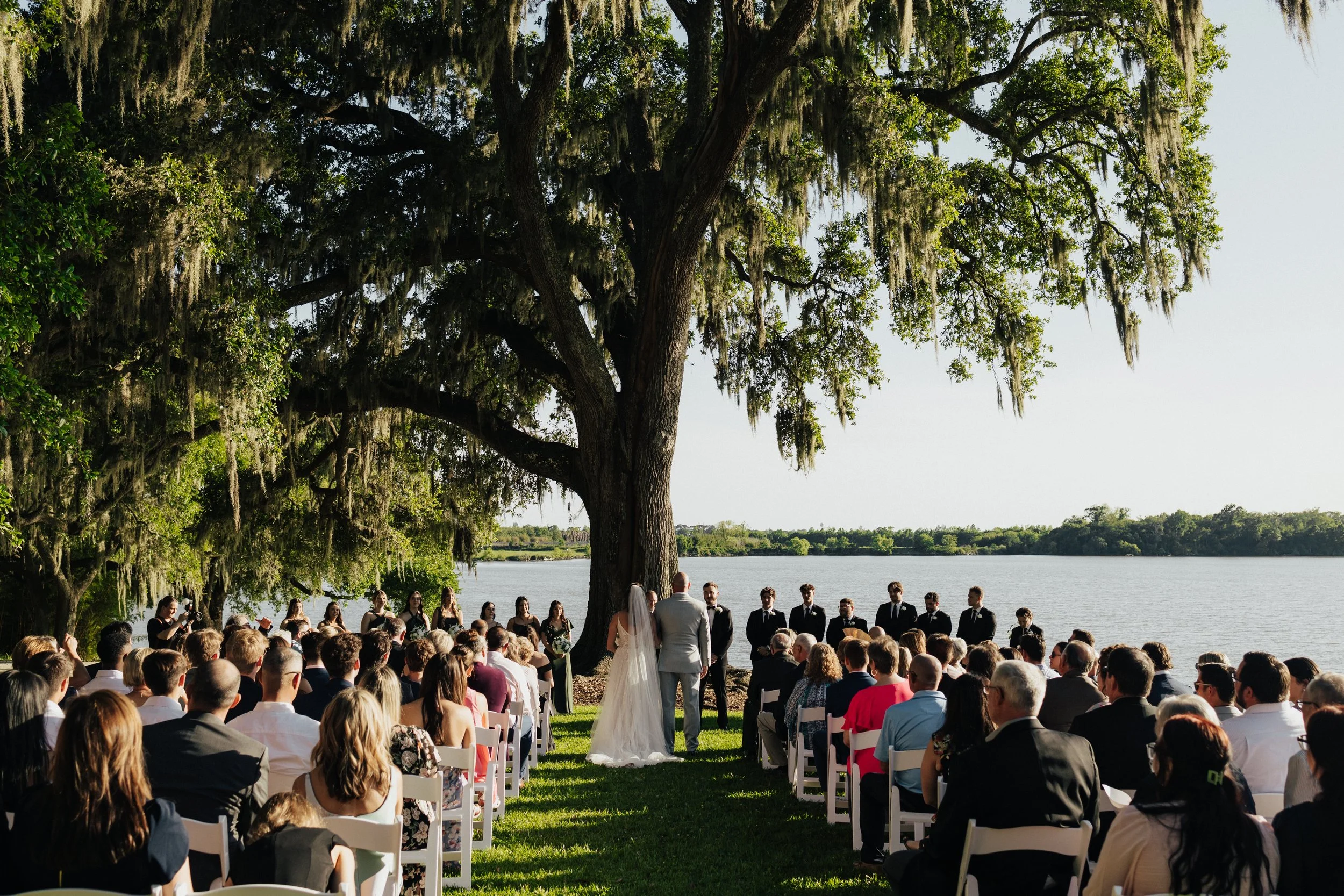 A large outdoor wedding ceremony by a lake with many guests seated on white chairs, a bride and groom standing in front of the officiant, a large tree with hanging moss overhead, and a scenic view of water and distant trees.