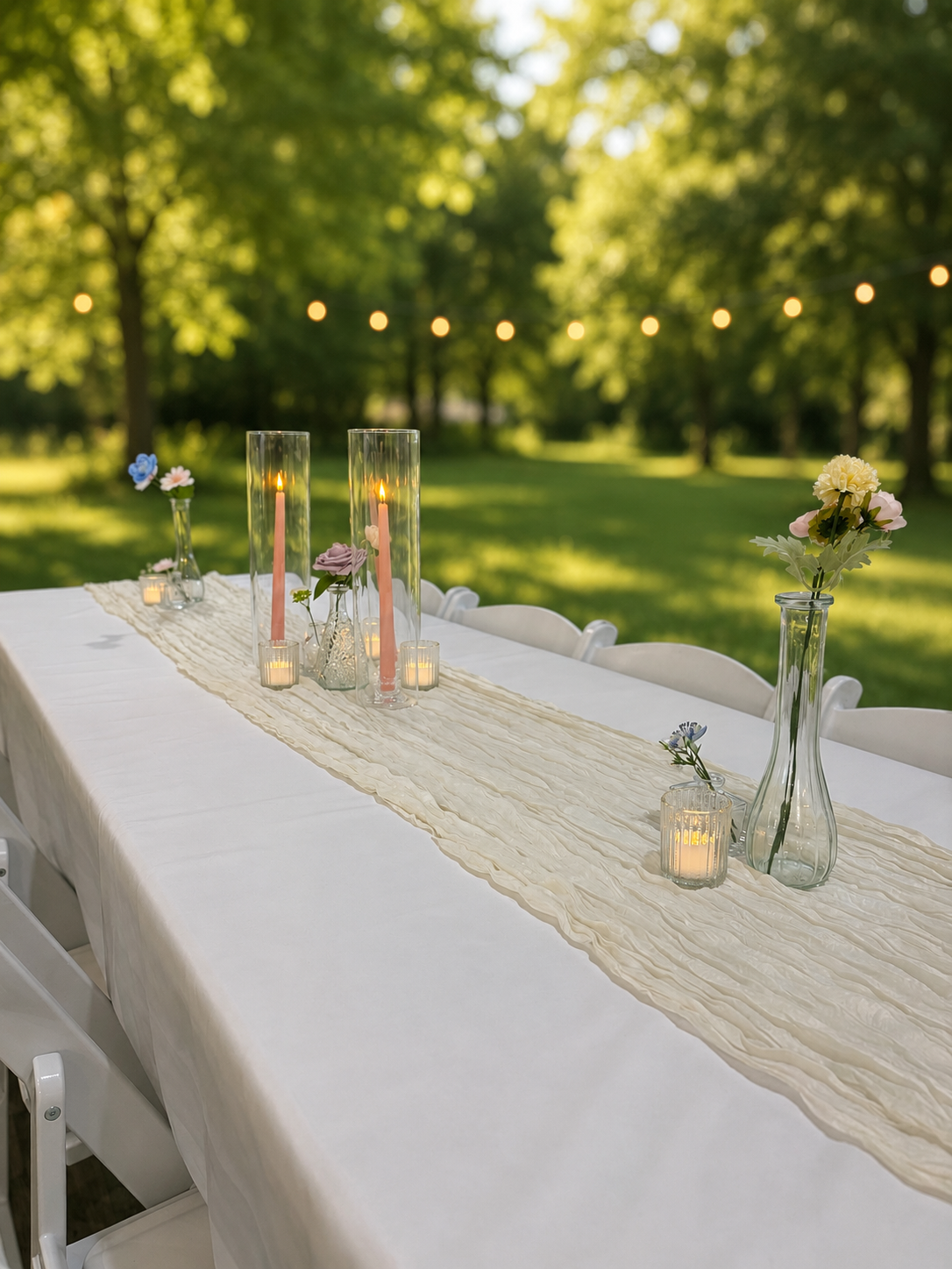 Decorated outdoor dining table with vases, candles, and flowers on a cream table runner in a garden setting with string lights overhead.