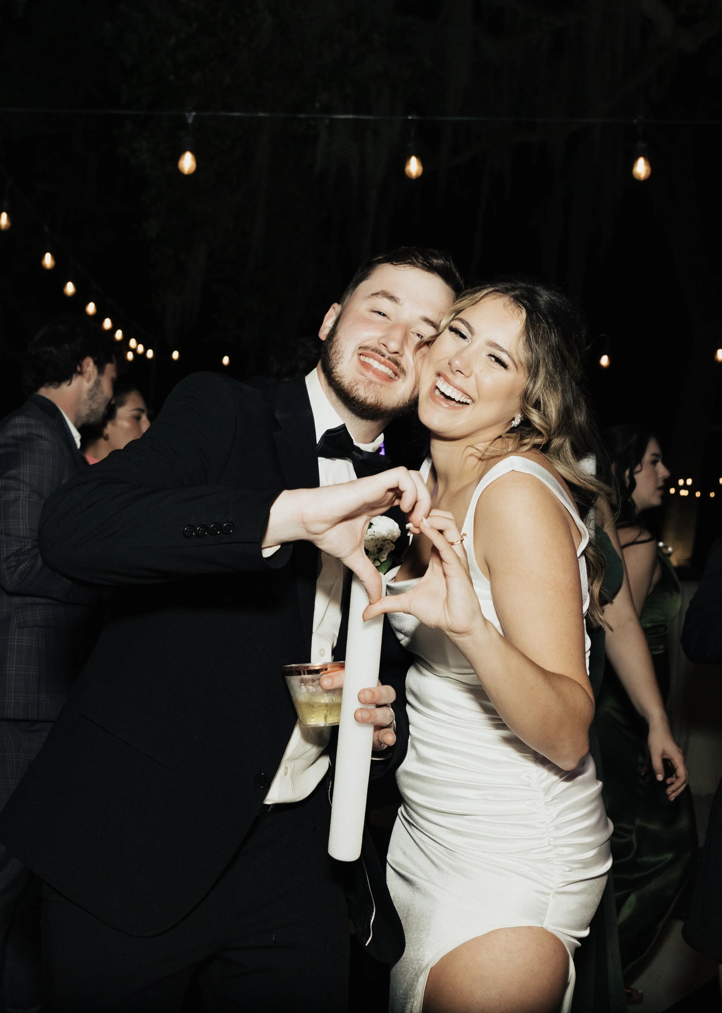 A smiling man in a tuxedo and a woman in a white dress are posing together at a celebration, forming a heart with their hands. They are surrounded by other guests at an event with string lights overhead at night.