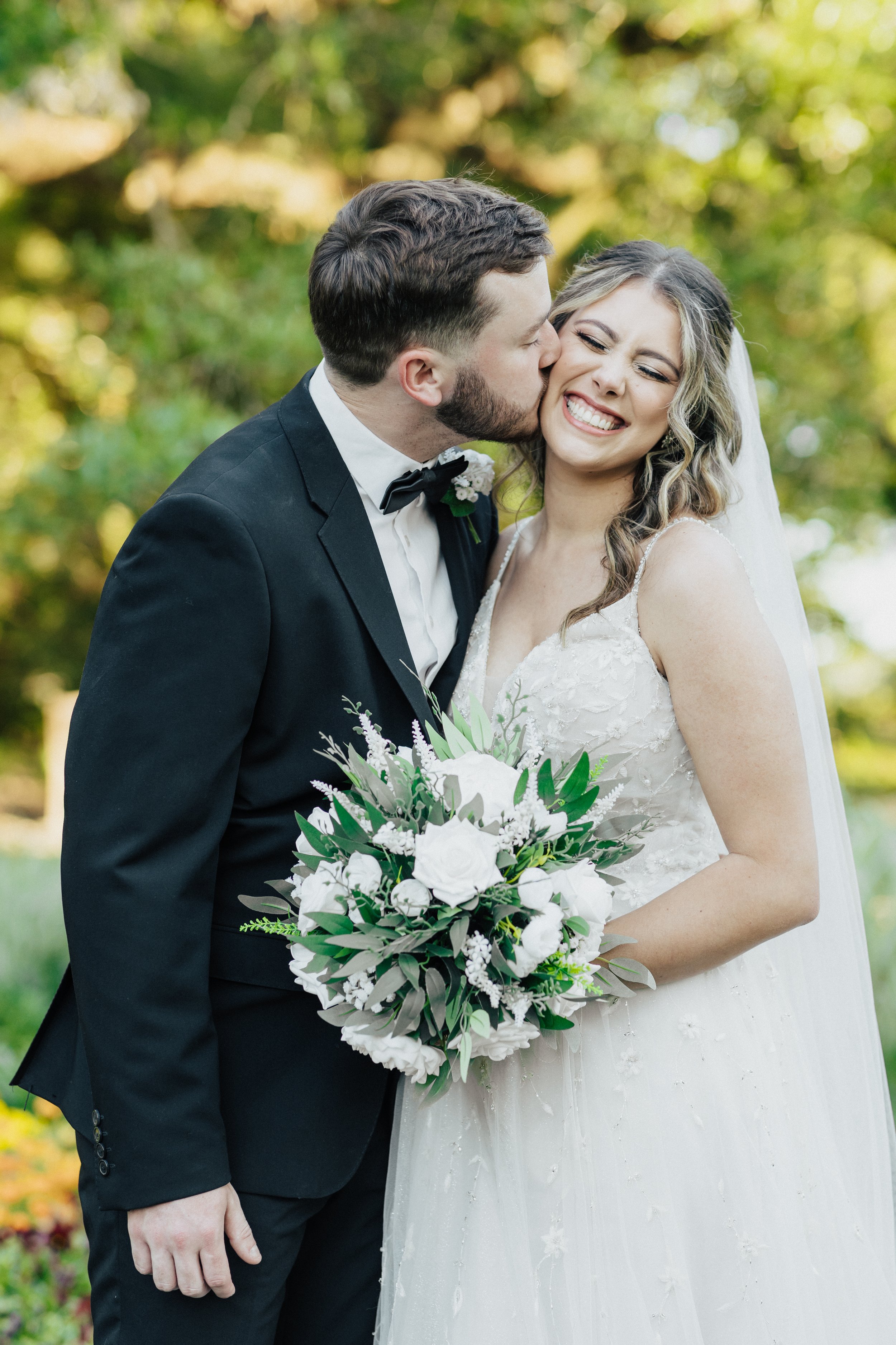 A newlywed couple outdoors during daytime; the groom, with dark hair and a beard, is kissing the bride on her cheek, who is smiling joyfully. The bride has blonde, wavy hair and is wearing a white wedding dress with lace details, holding a bouquet of white flowers and greenery.