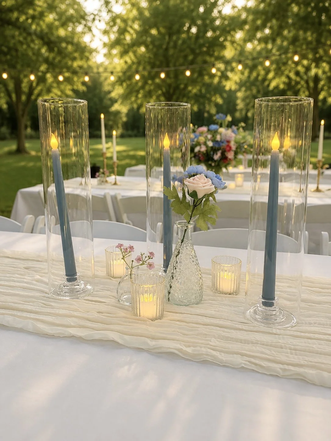 A decorated outdoor table with candles, flowers, and glass vases, set for an event in a lush green garden during sunset.