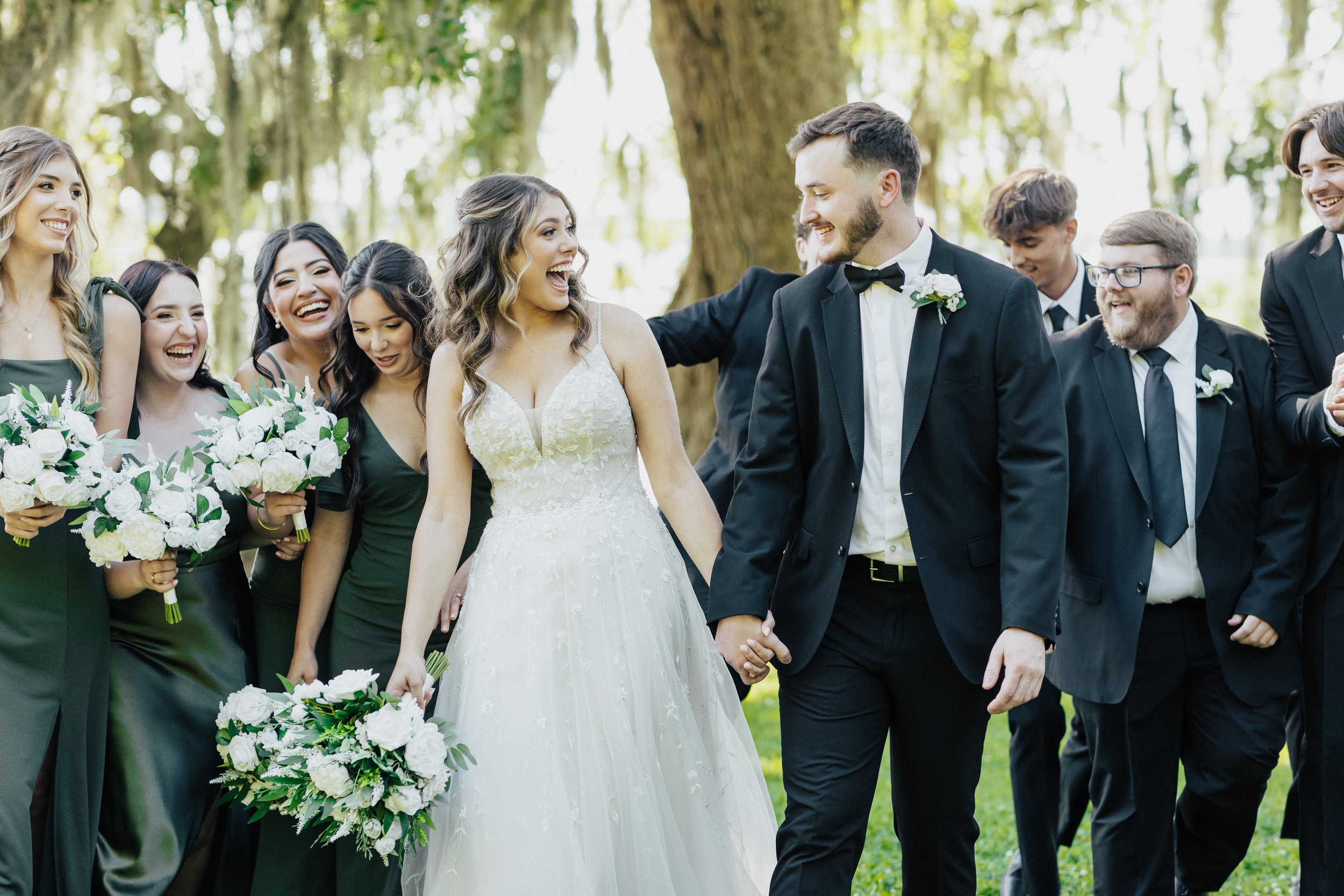 A wedding scene featuring the bride and groom holding hands, surrounded by friends in formal attire outdoors in a wooded area, with trees and green foliage in the background.