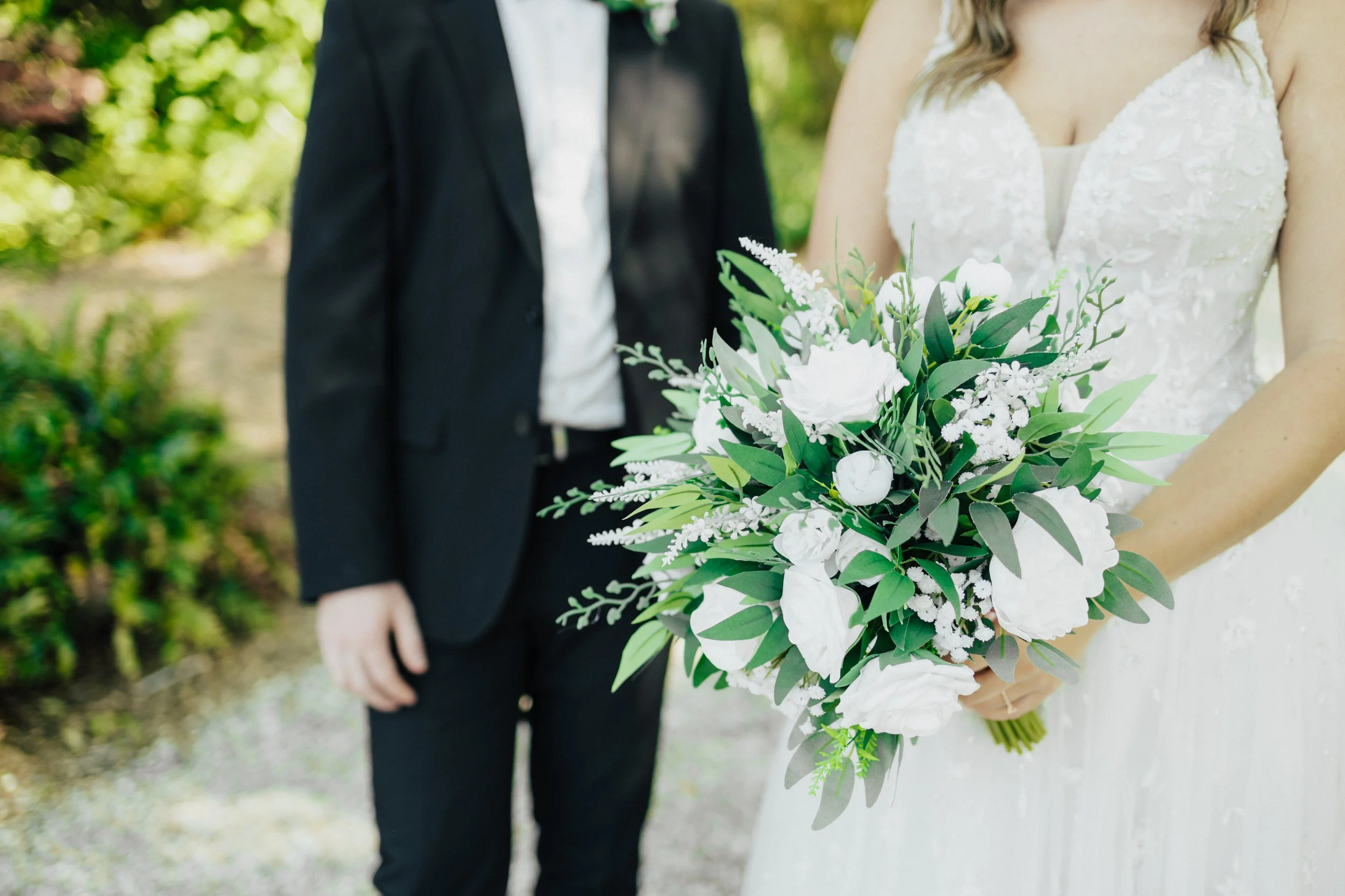 Bride holding a bouquet of white flowers and greenery, standing next to a groom in a black tuxedo outdoors