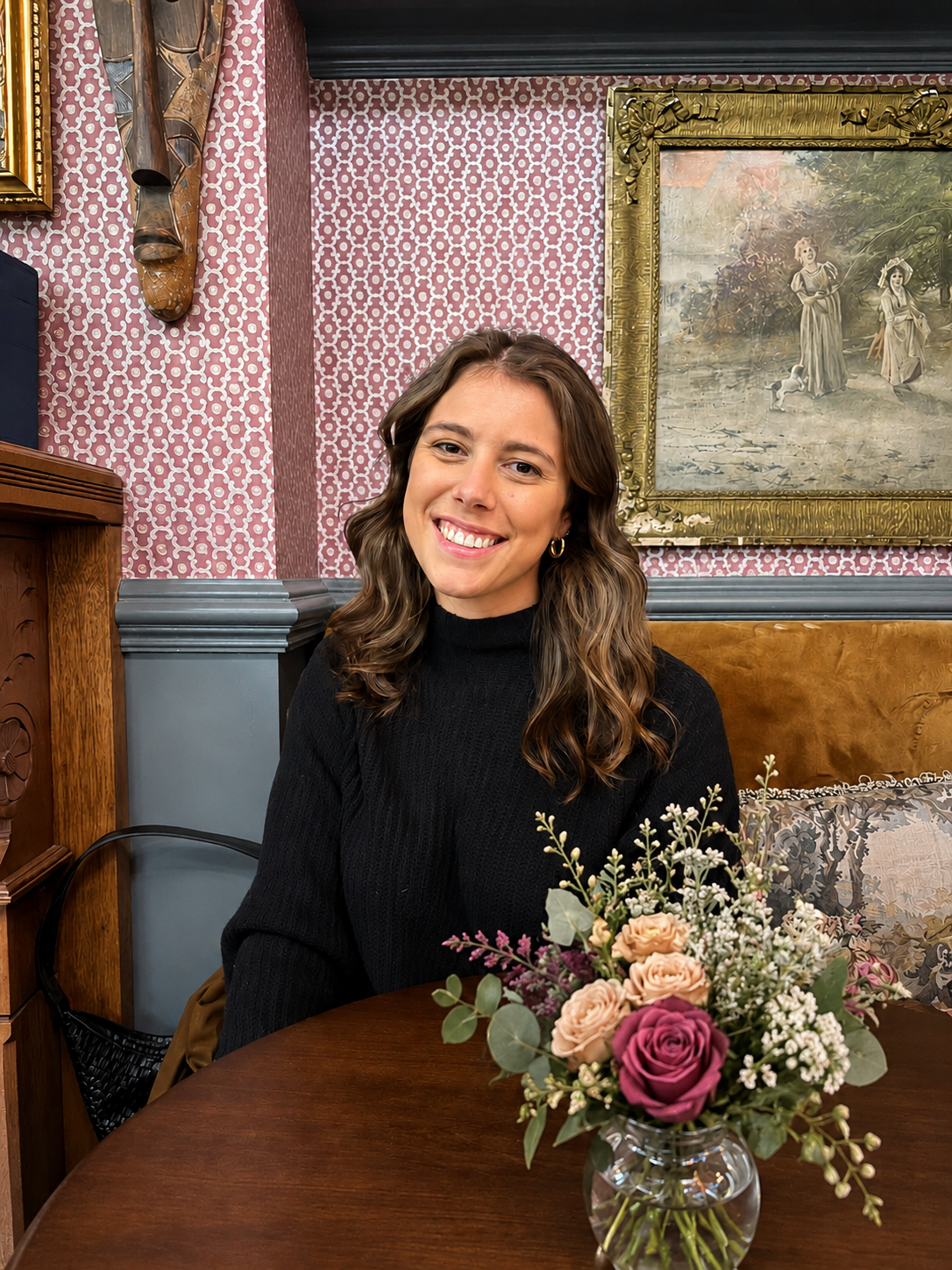 A young woman with shoulder-length curly brown hair, wearing a black sweater, sitting at a round wooden table with a floral arrangement in a glass vase. The background features patterned pink wallpaper, a gold-framed painting, and decorative wooden wall art.