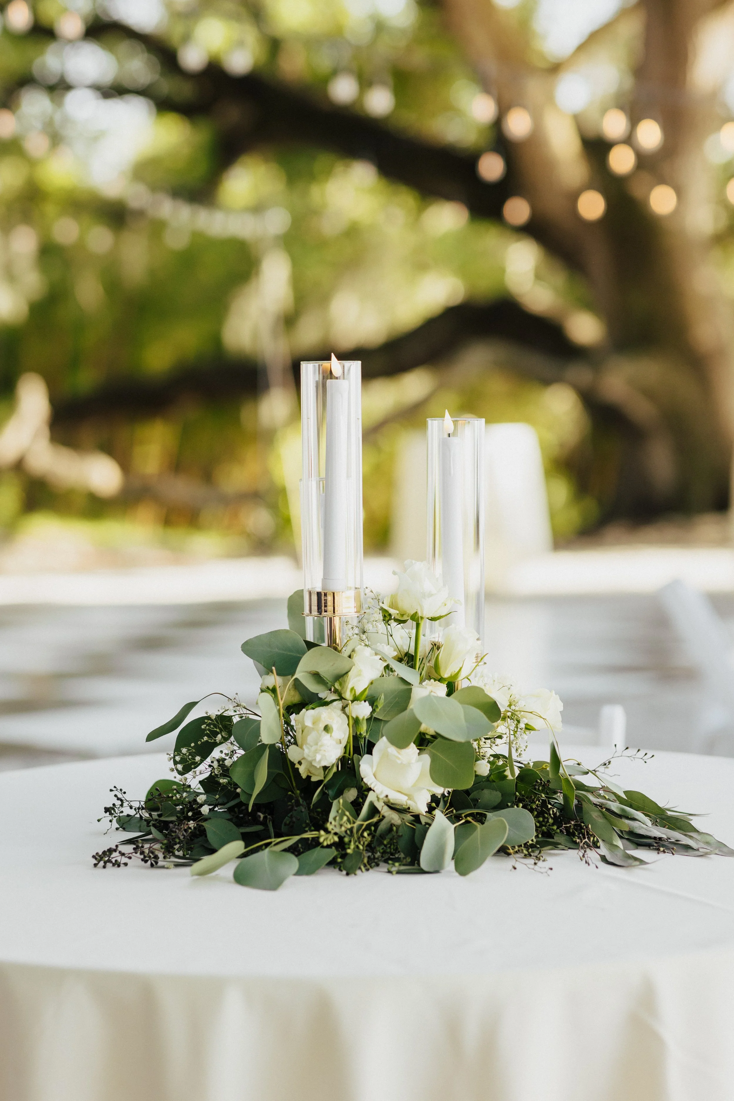 Close-up of a floral centerpiece with white flowers and green foliage on a white table, with blurred outdoor background.