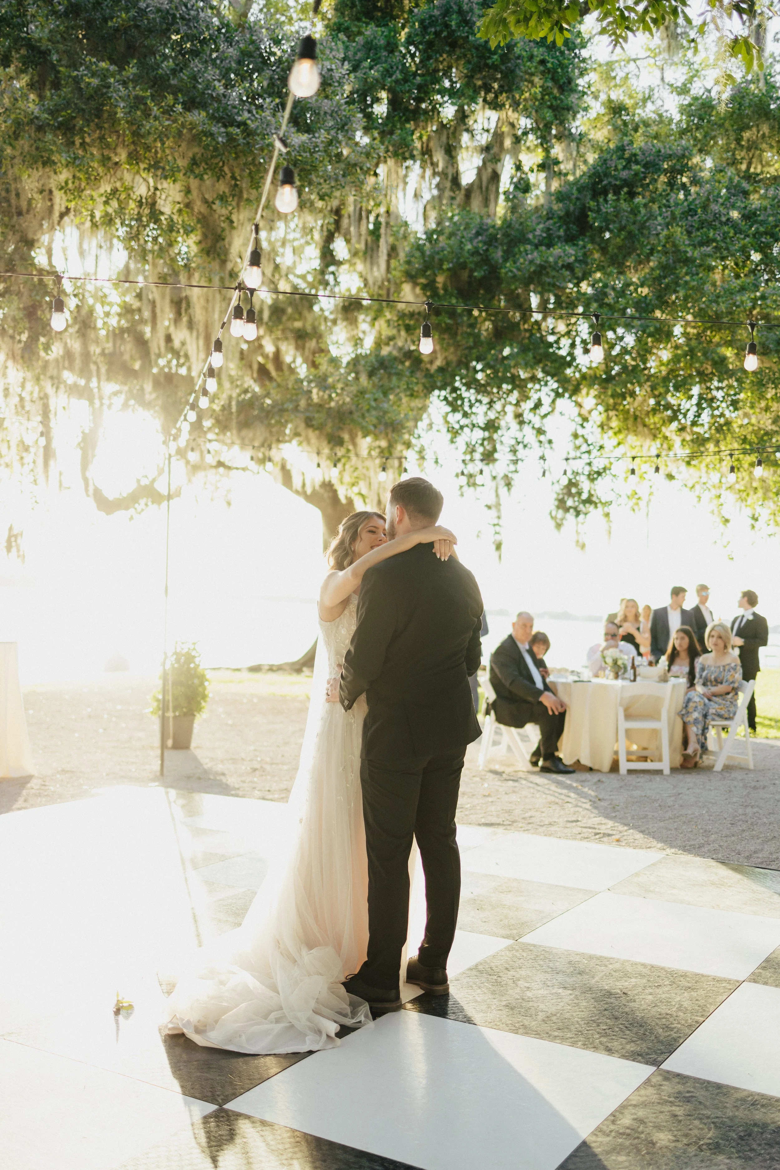 A bride and groom dancing together at their outdoor wedding reception during sunset, surrounded by seated guests and string lights hanging overhead.