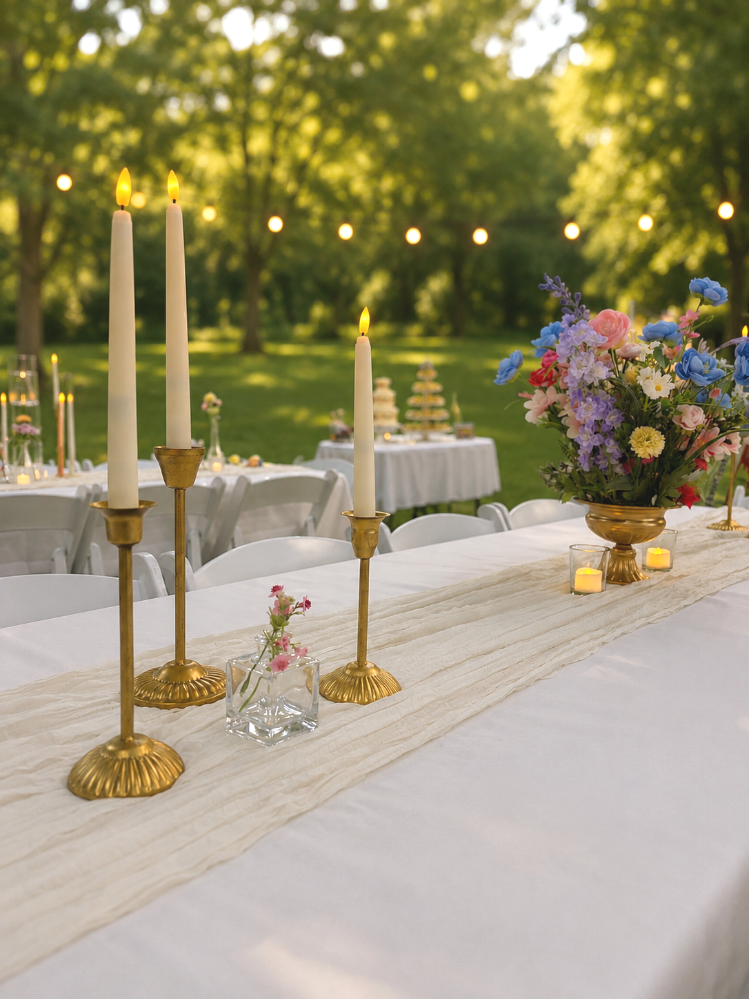 Outdoor wedding reception table with gold candleholders, tall white candles, a flower arrangement with pastel and purple flowers, and small candles, set on a white tablecloth with a cream-colored runner, surrounded by white chairs, with a background of trees and string lights.