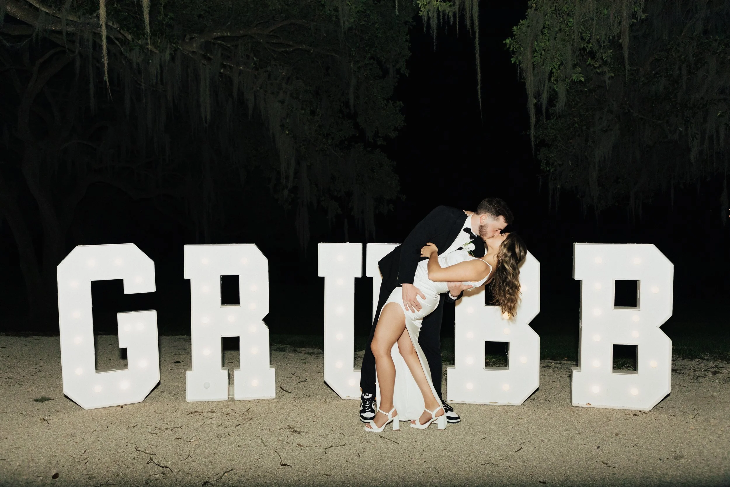A newlywed couple kissing in front of large illuminated letters spelling 'LOVE' at night, with dark trees in the background and the bride wearing a white dress and high heels, and the groom in a black tuxedo.