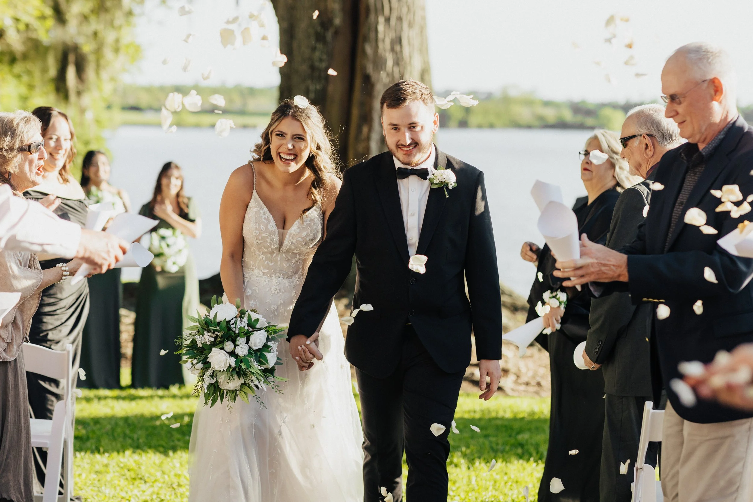A bride and groom holding hands during their outdoor wedding ceremony on a sunny day, surrounded by guests throwing flower petals.