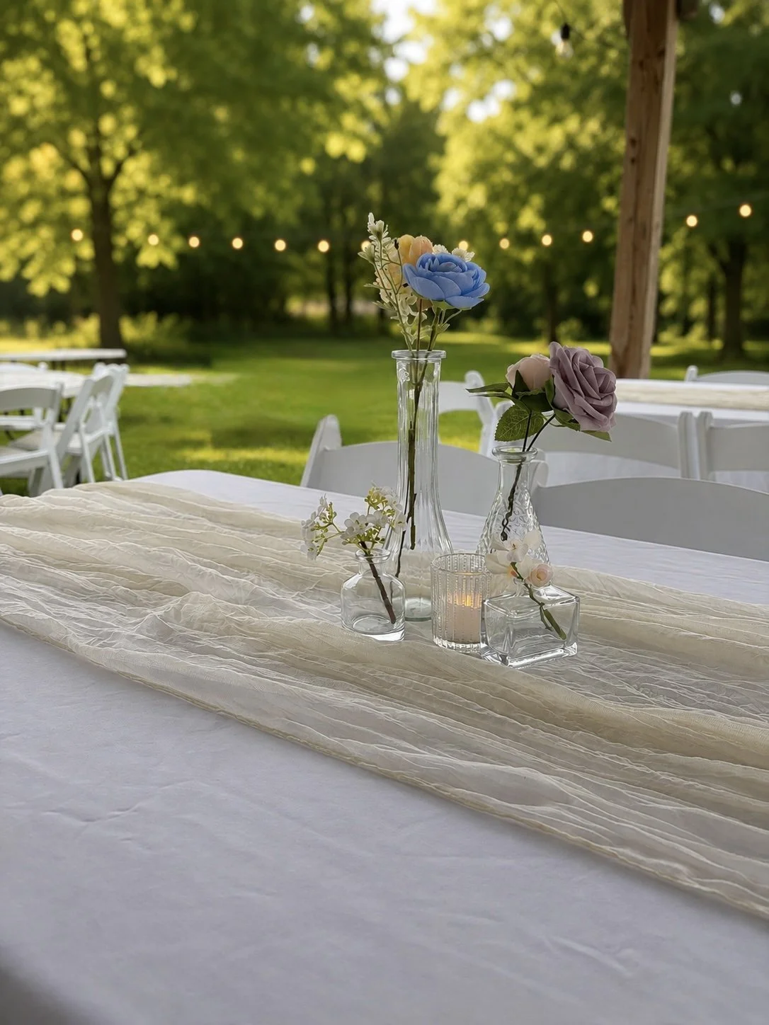 Table decorated with glass vases and flowers, set outdoors with grass and trees in the background, and string lights overhead.