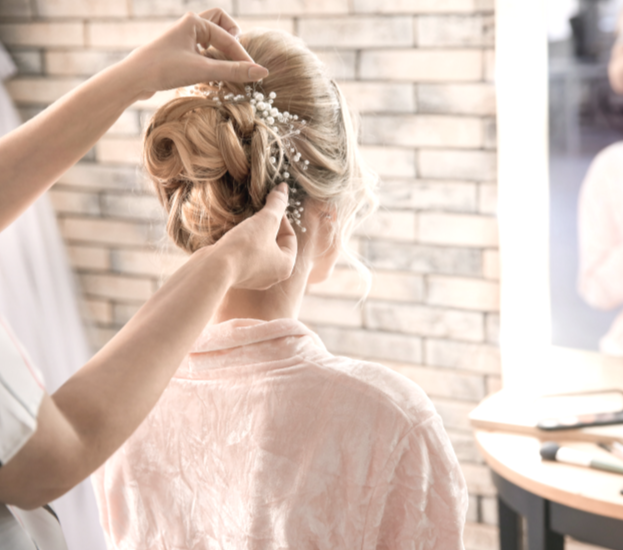 A woman with blonde hair styled in an elegant updo is having a decorative hair accessory arranged with small white flowers and pearl accents, by a hairstylist in a salon or studio with brick wall background.