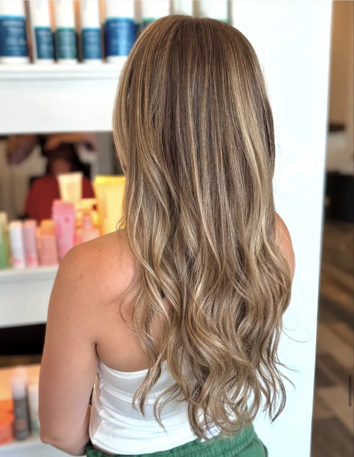 Back view of a woman with long, curled, blonde hair in a salon setting with shelves of hair products in the background.